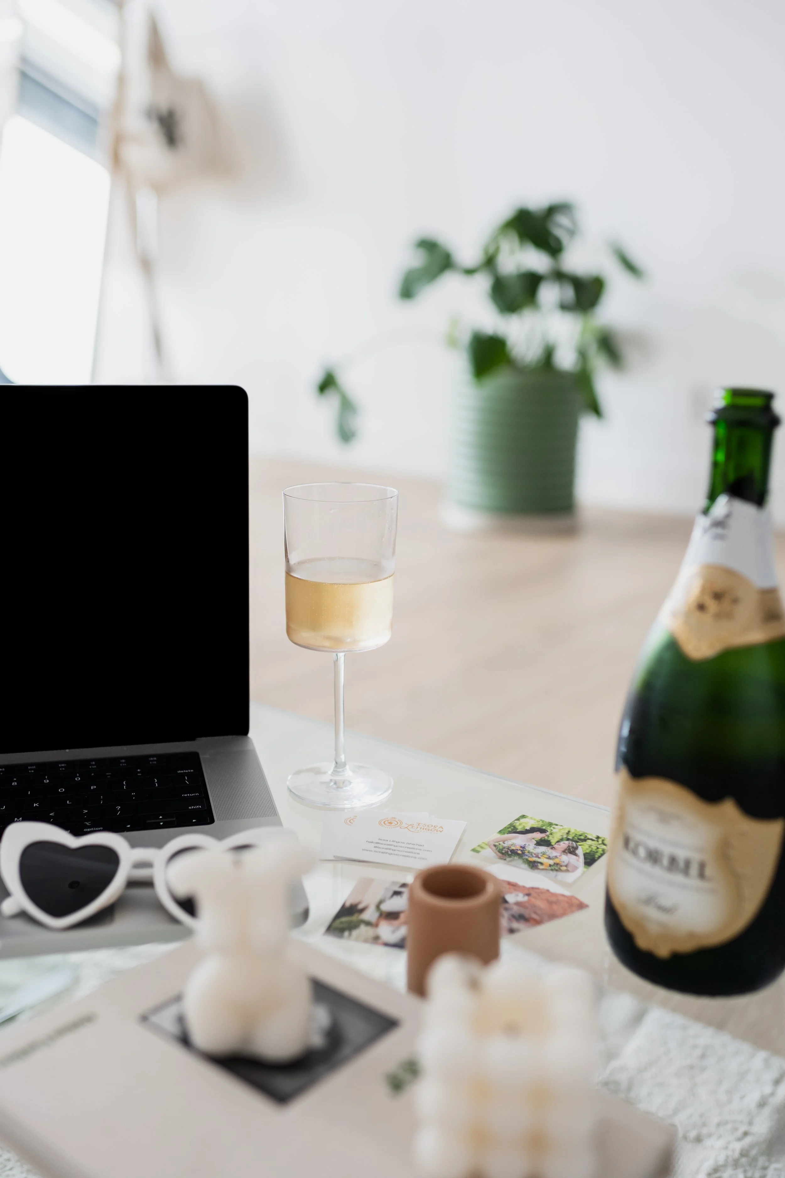 A workspace with a laptop, a glass of white wine, a bottle of champagne, decorative candles, heart-shaped glasses, and some photographs on a white table, with a potted plant and a window in the background.