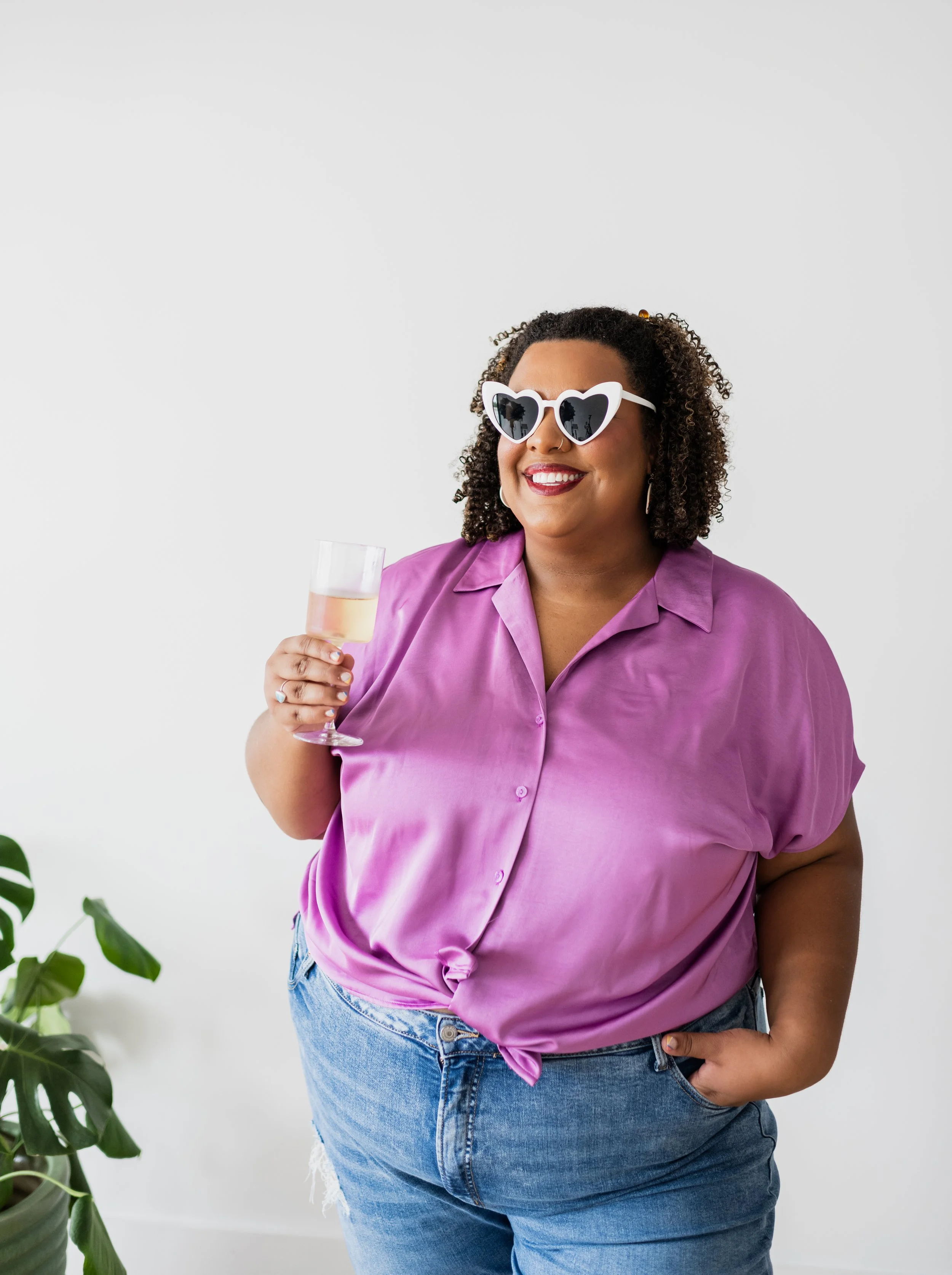 A cheerful woman with curly hair wearing heart-shaped sunglasses and a purple satin shirt, holding a glass of champagne, standing with her hand in her pocket against a plain white background.