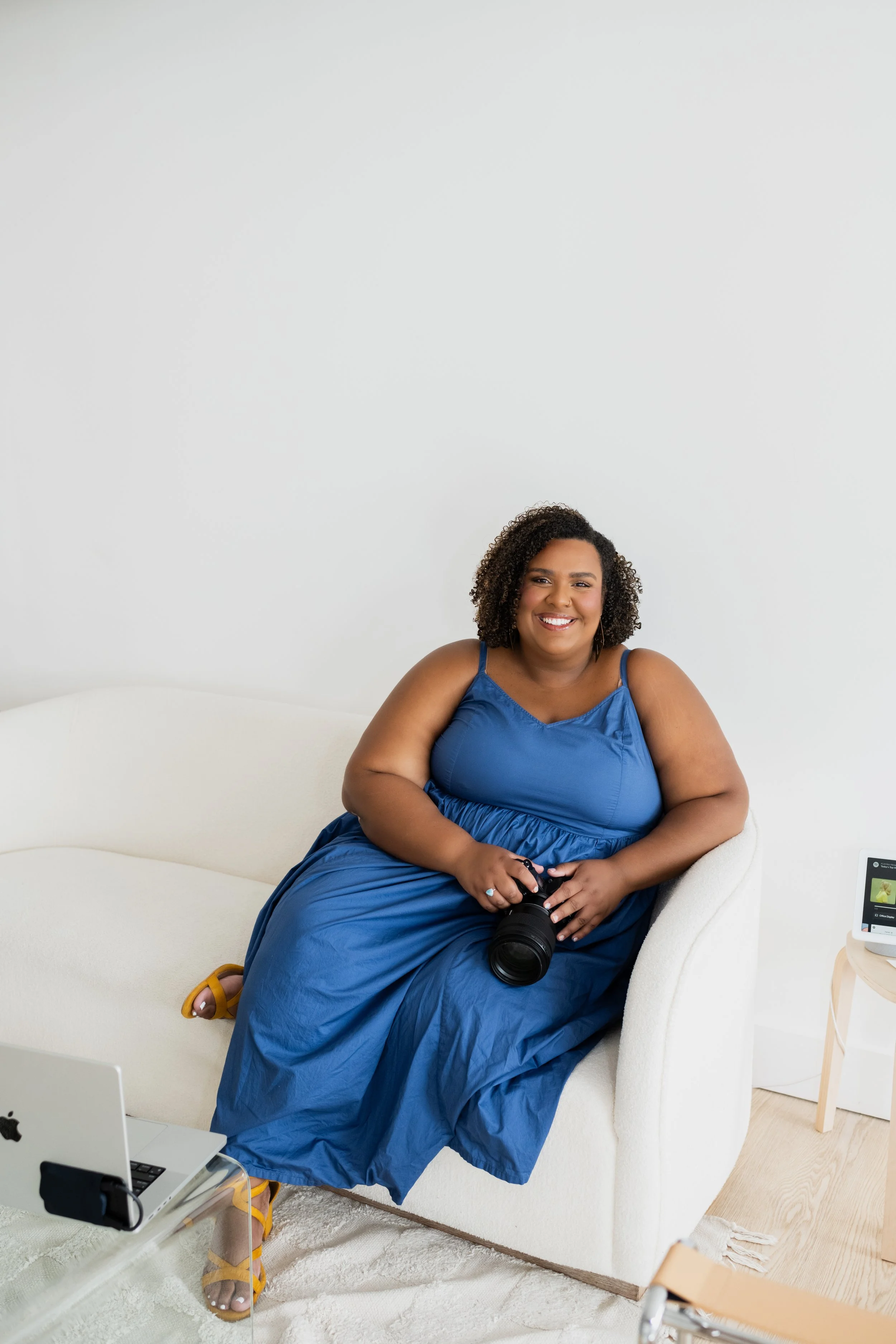 Smiling woman in a blue dress sitting on a white couch, holding a camera, in a bright room with a laptop, tablet, and white walls.