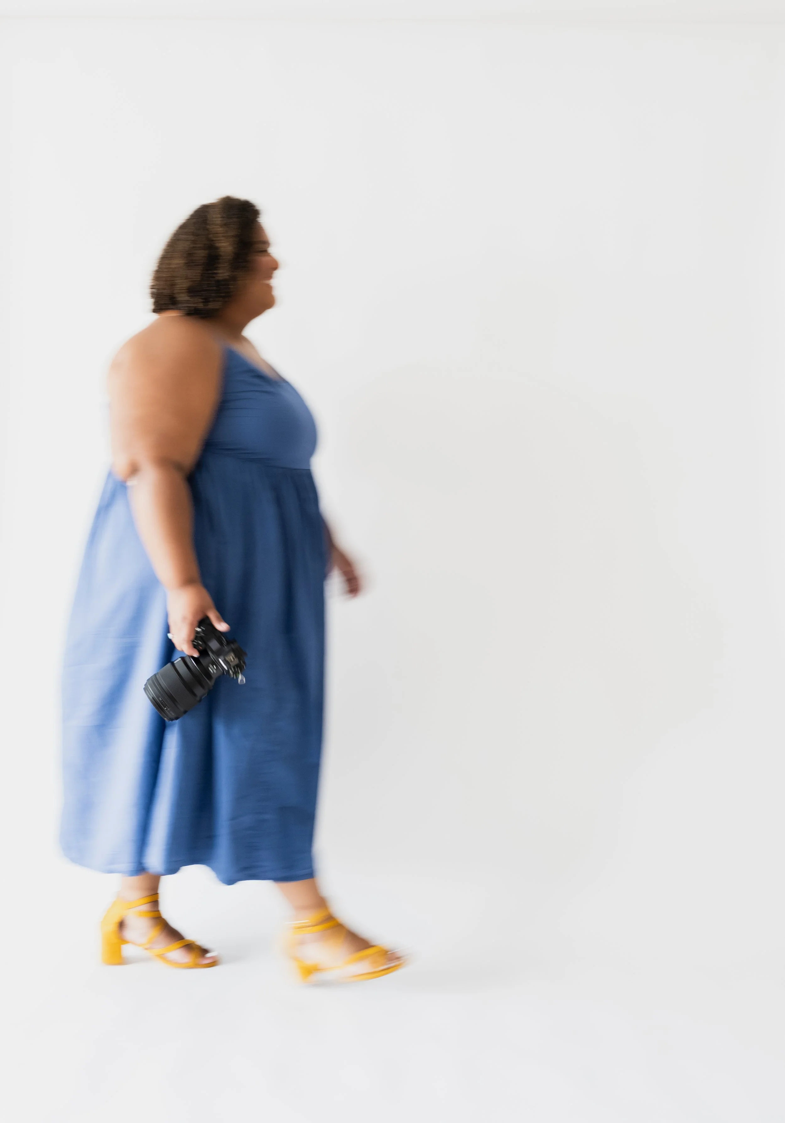 A woman walking in a blue dress and yellow heels, holding a camera, against a plain white background.