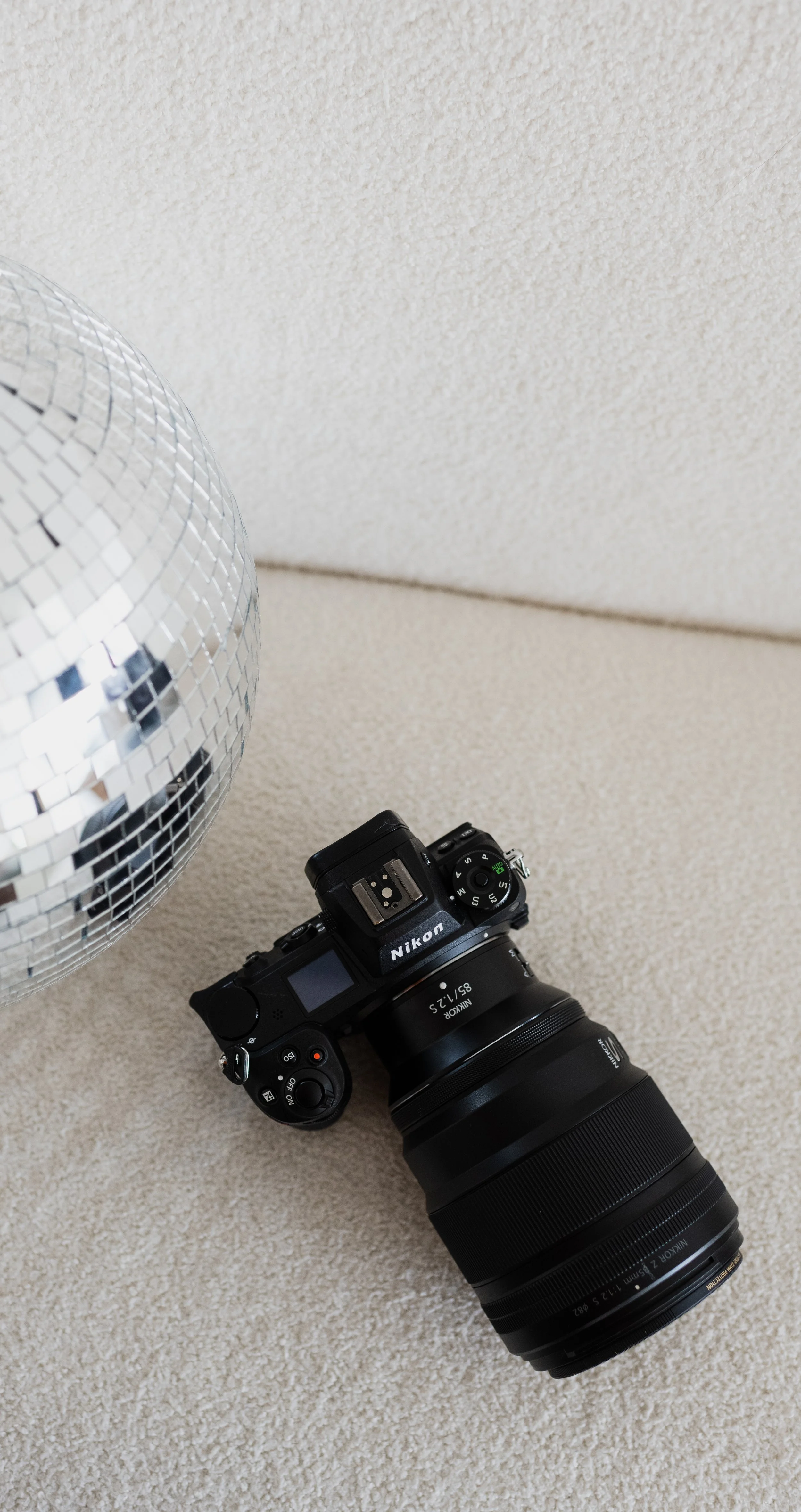 A black Nikon DSLR camera with a zoom lens placed on a beige carpeted floor next to a large mirror ball.