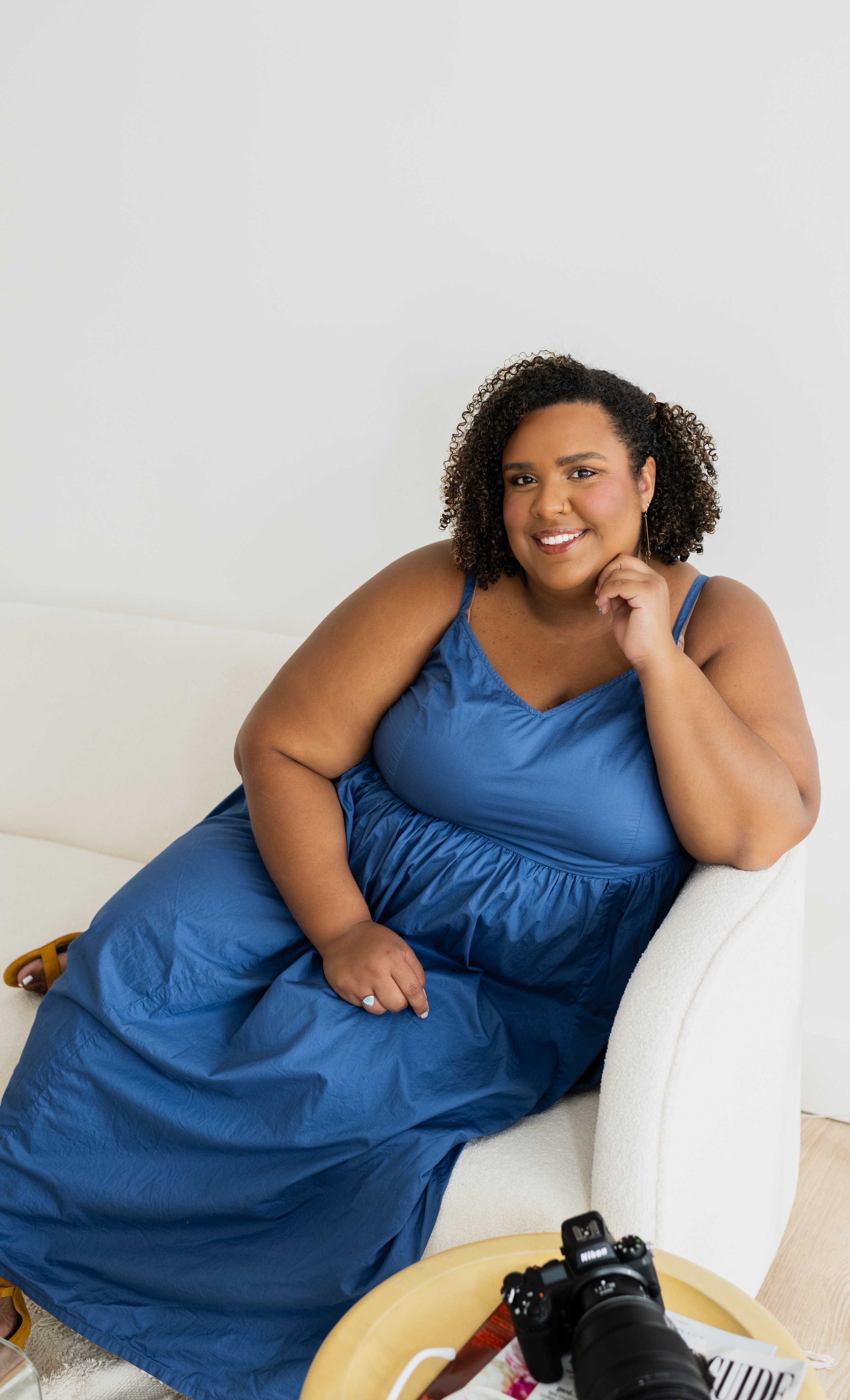 A woman with curly hair and a blue dress sitting on a white couch, smiling while resting her chin on her hand. A camera is on a small table in front of her.
