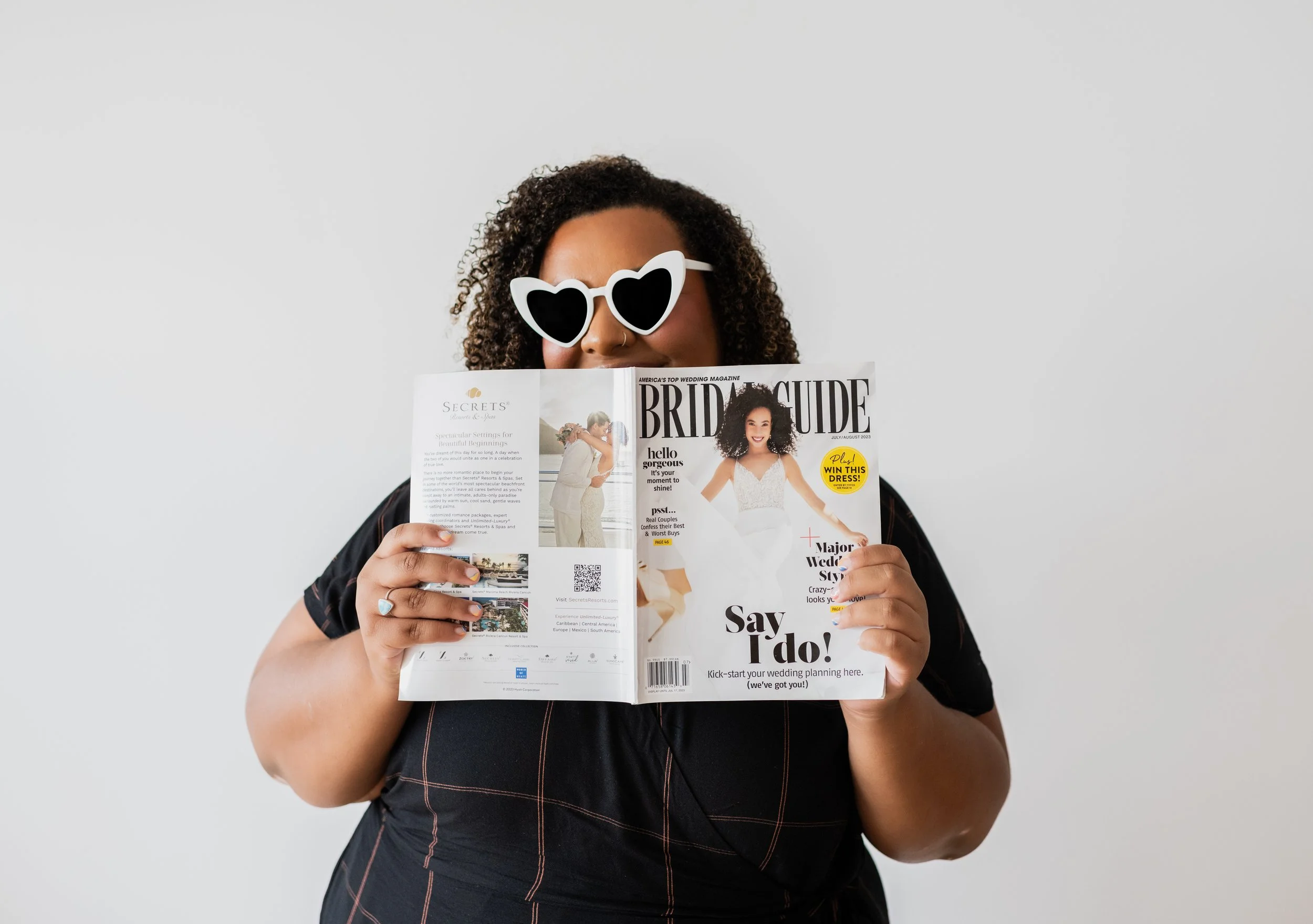 A woman with curly hair wearing white heart-shaped sunglasses holding a wedding magazine in front of her face against a plain light gray background.