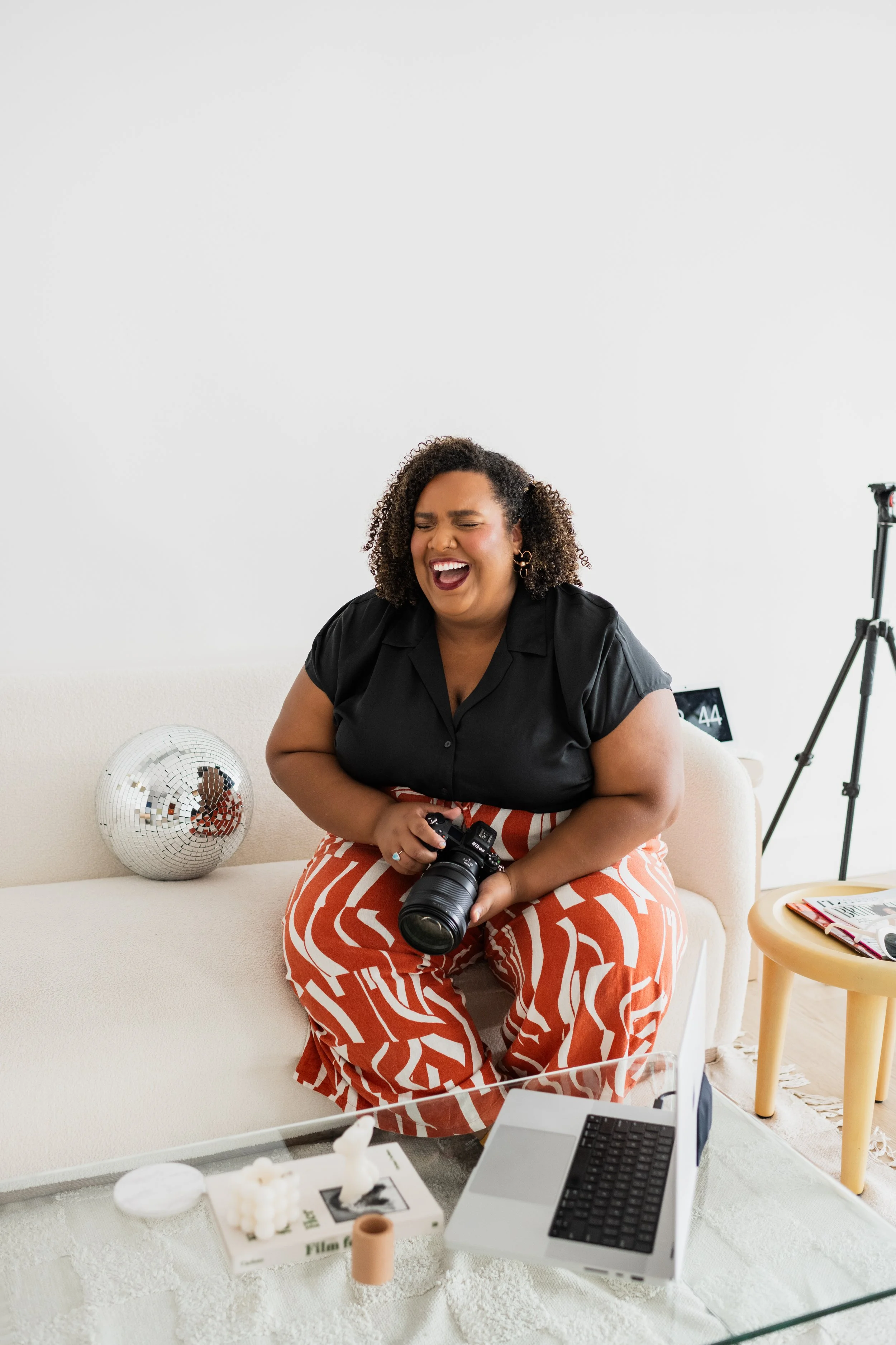 Woman with curly hair sitting on a white sofa, holding a camera and laughing. A disco ball is on the sofa beside her. There is a laptop, magazines, and decorative items on a glass coffee table in front of her.
