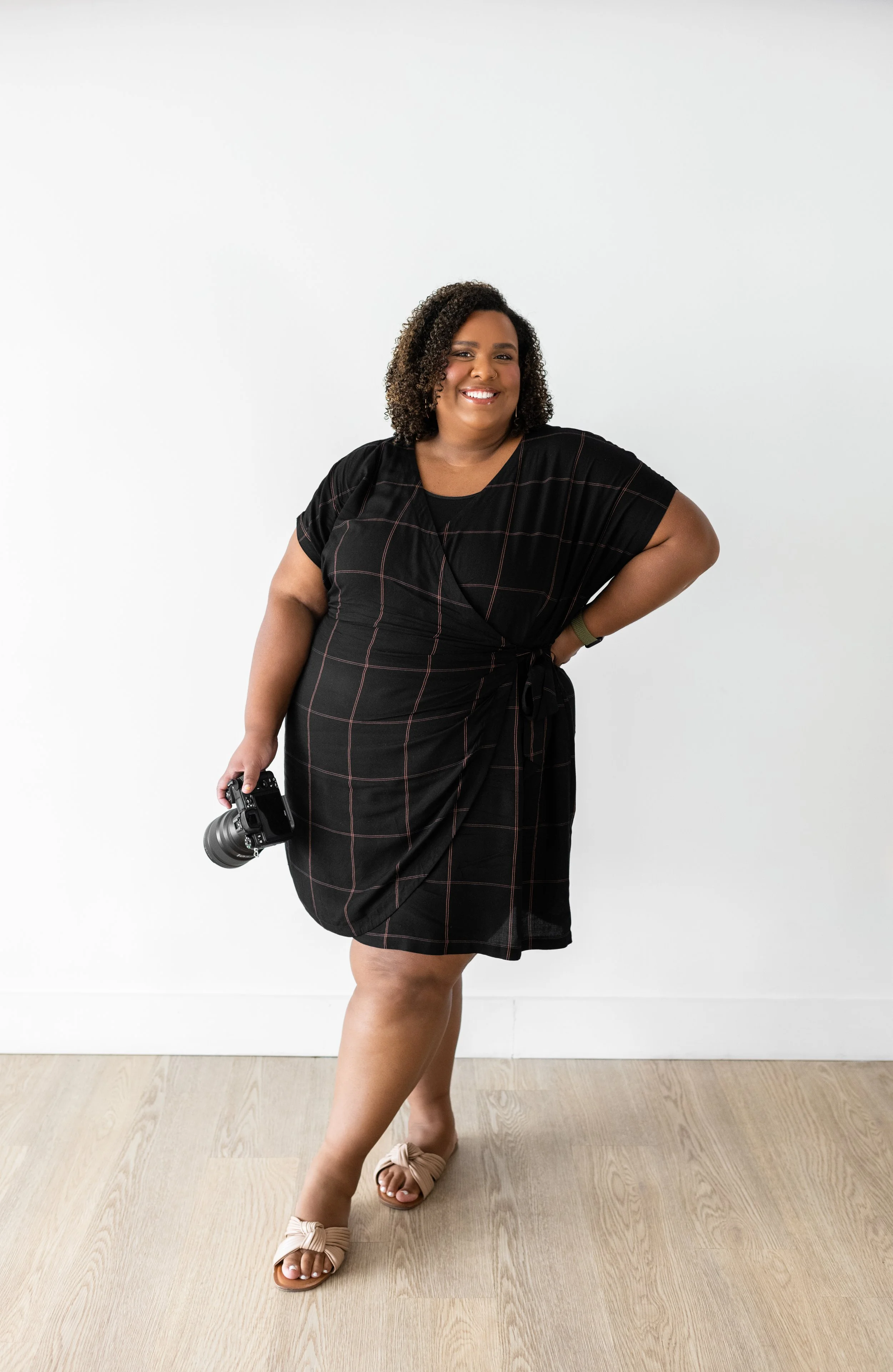 A woman with curly hair wearing a black dress with a windowpane pattern, beige sandals, and holding a camera, standing against a white wall, smiling.