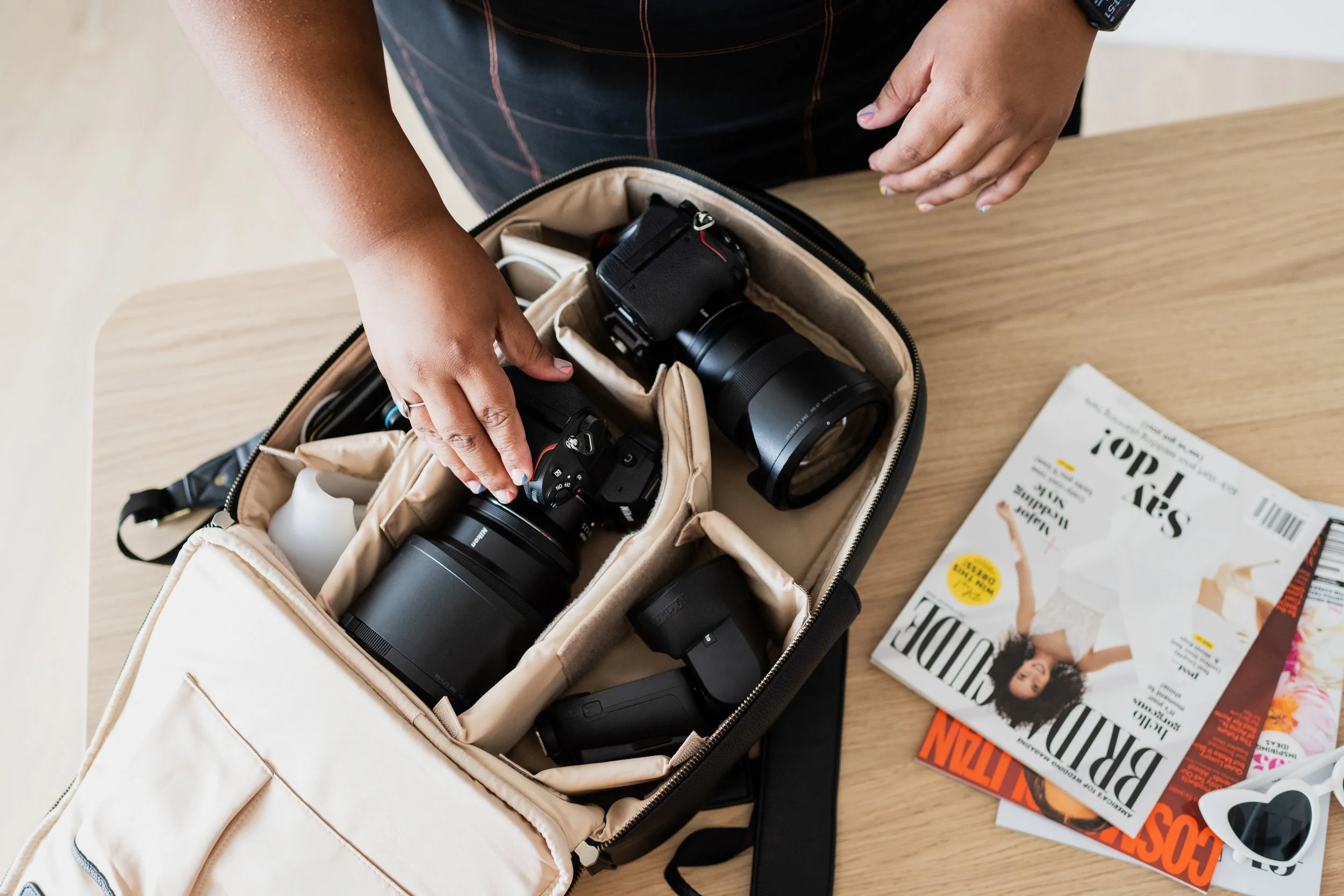 Person packing camera equipment into a beige camera bag on a wooden table, with magazines and sunglasses nearby.