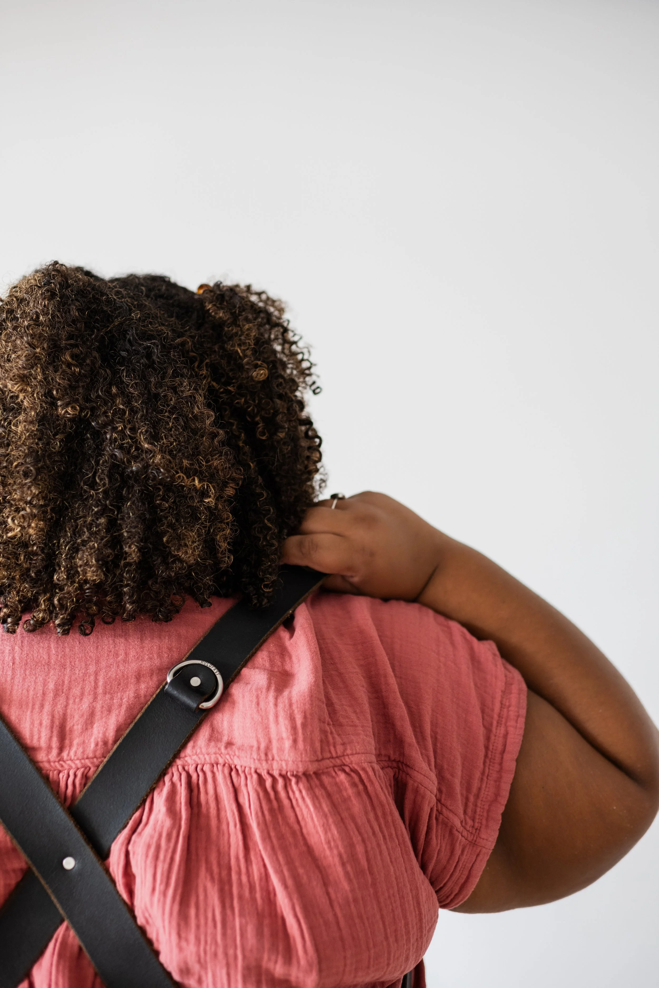 A woman with curly dark hair and a pink top is adjusting a black strap on her shoulder against a plain white wall.