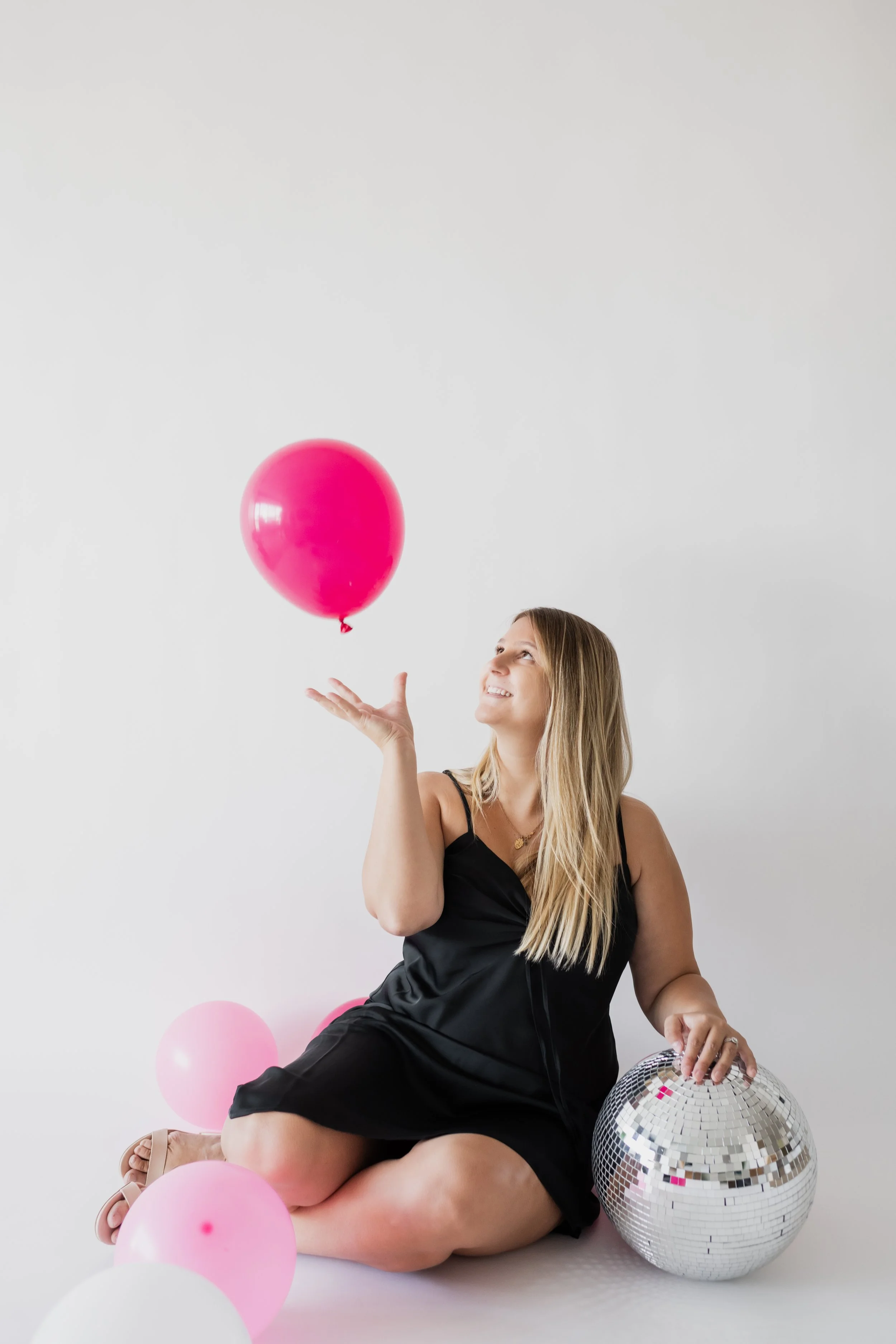 A woman sitting on the floor, wearing a black dress, smiling and tossing a pink balloon in the air next to a disco ball, with pink and white balloons around her, against a plain white background.