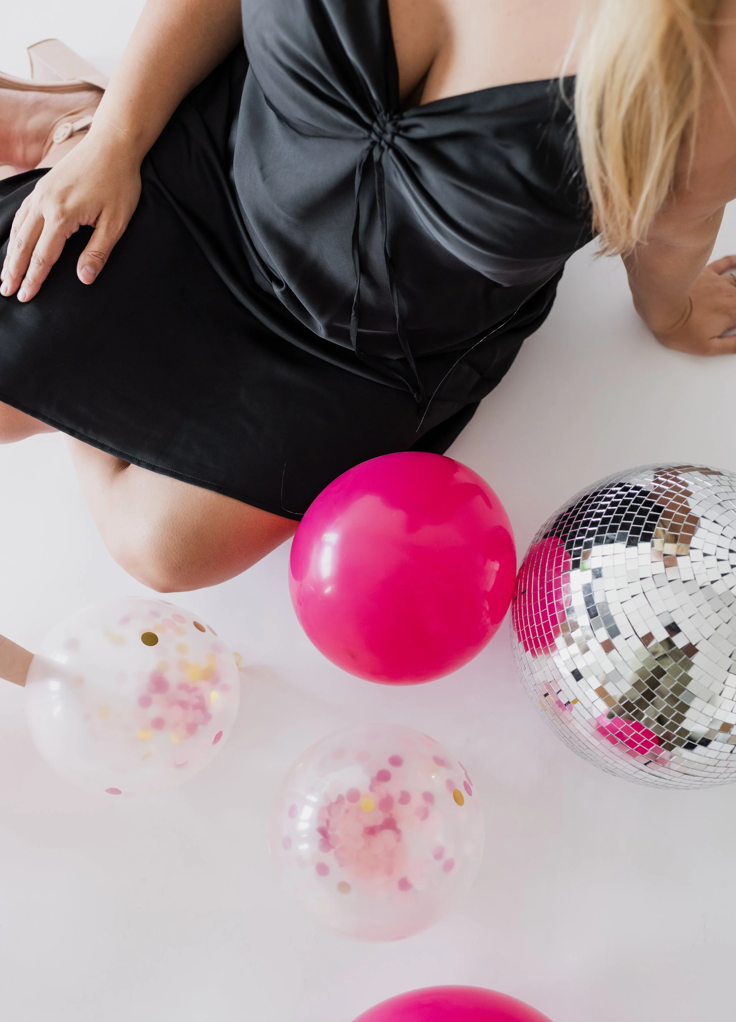 A woman in a black satin dress sitting on a white surface surrounded by pink, clear with confetti, and silver mirror disco balloons.