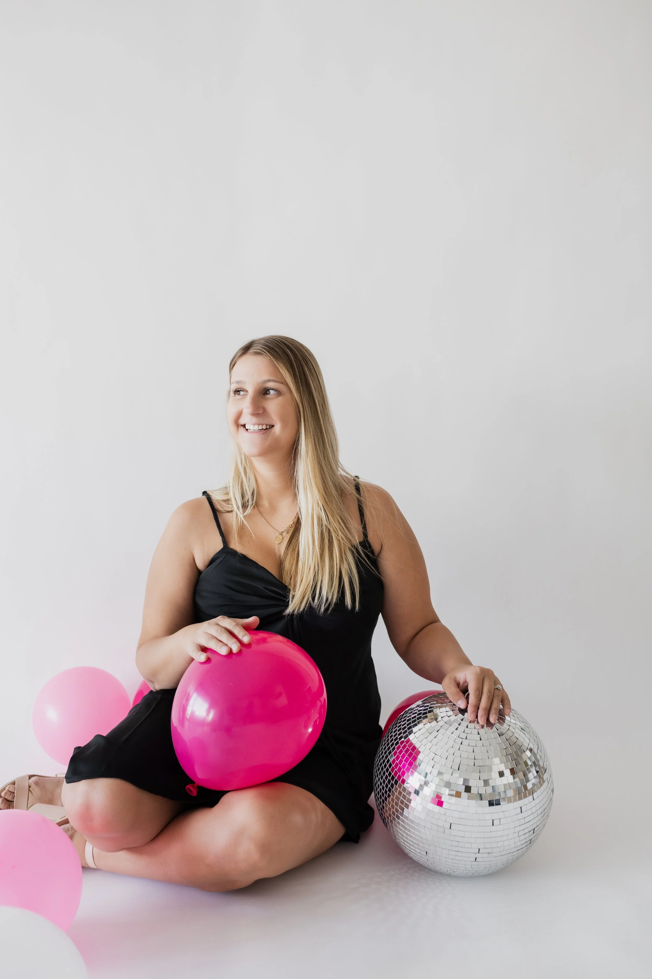 A woman sitting on the floor surrounded by pink and white balloons and a large disco ball, smiling and looking to the side.