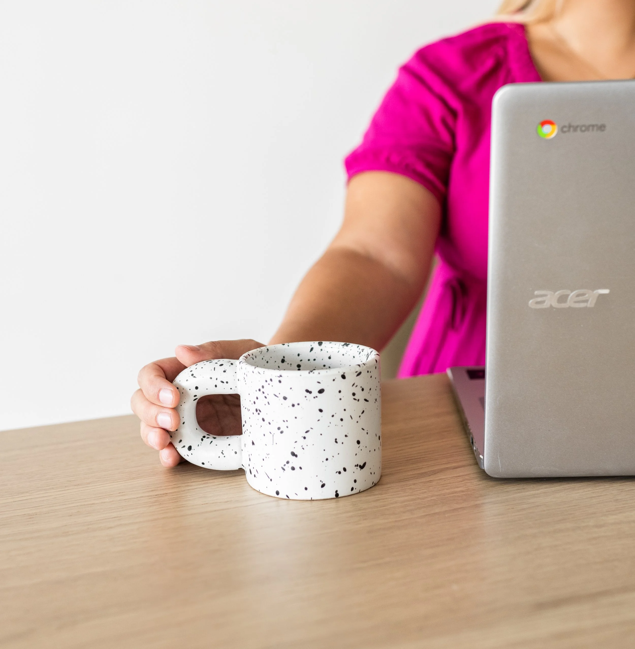 Person wearing a pink shirt holding a white speckled mug while sitting at a wooden table with a silver Acer laptop.