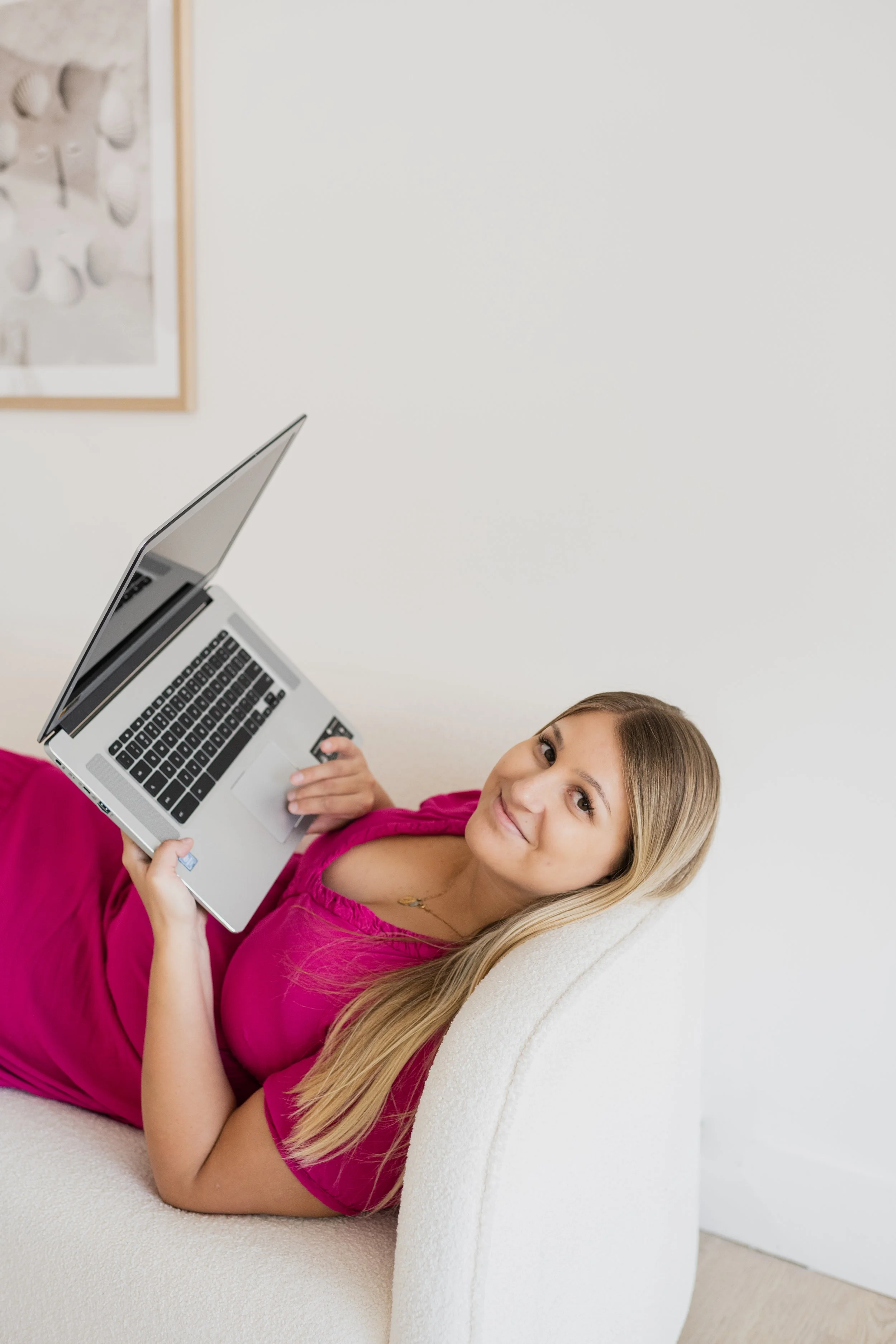 A young woman with long blonde hair lying on a cream-colored sofa, smiling, holding an open laptop with a black keyboard and silver body, wearing a bright pink dress, in a room with a white wall and a framed abstract art piece.