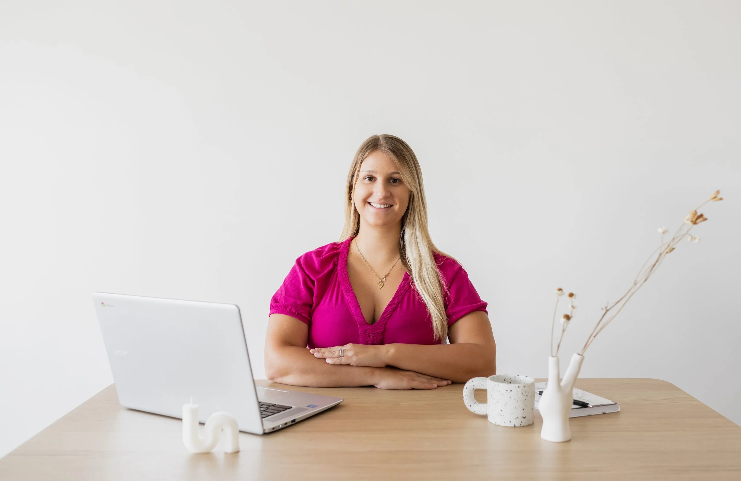 A woman with long blonde hair smiling and sitting at a desk with a silver laptop, white candle holder, and a white speckled coffee mug, with a white vase holding dried branches, against a plain white wall.