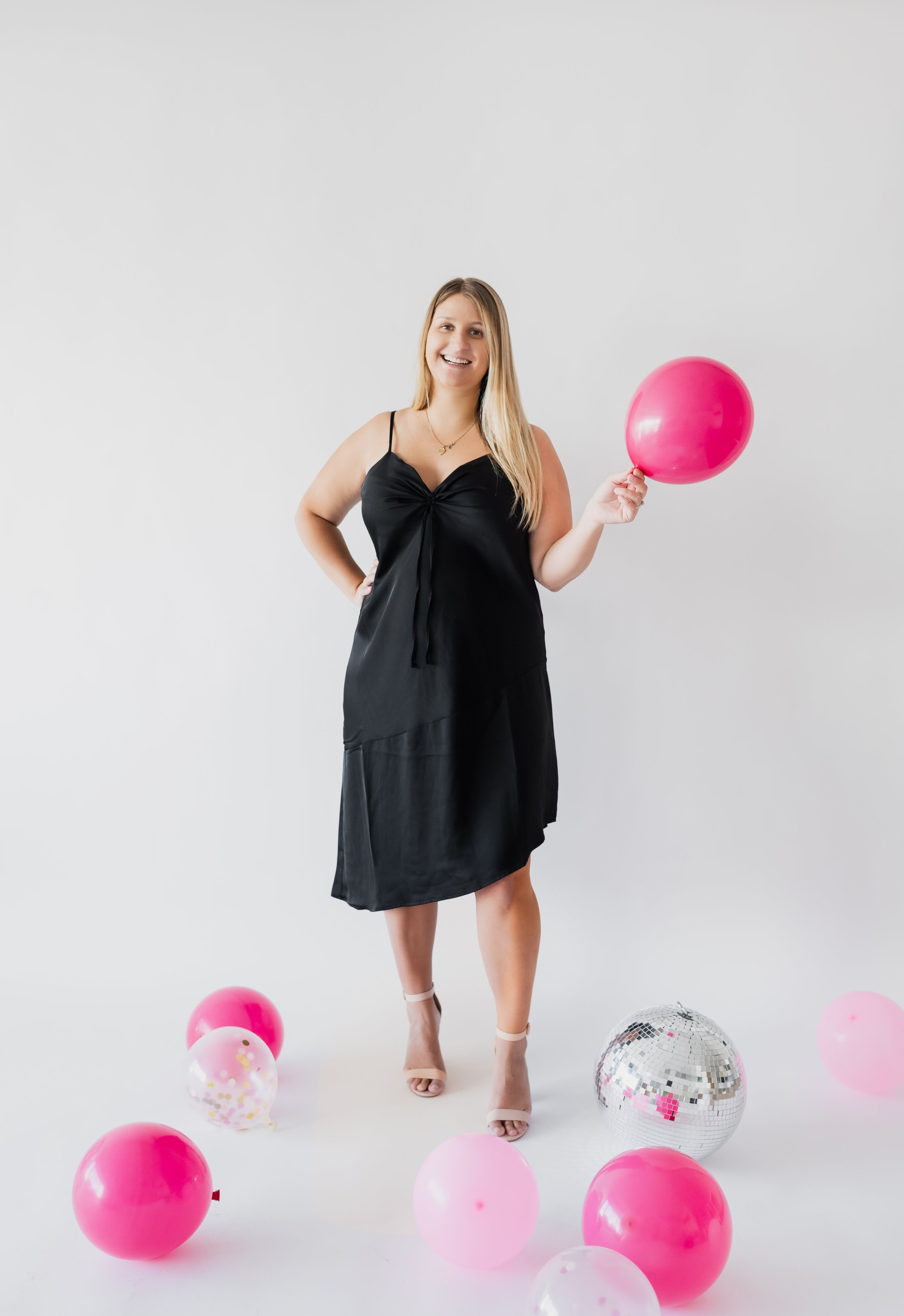 A woman with long blonde hair in a black dress holding a pink balloon, standing among pink and clear balloons, with a silver disco ball on the floor, against a plain white background.