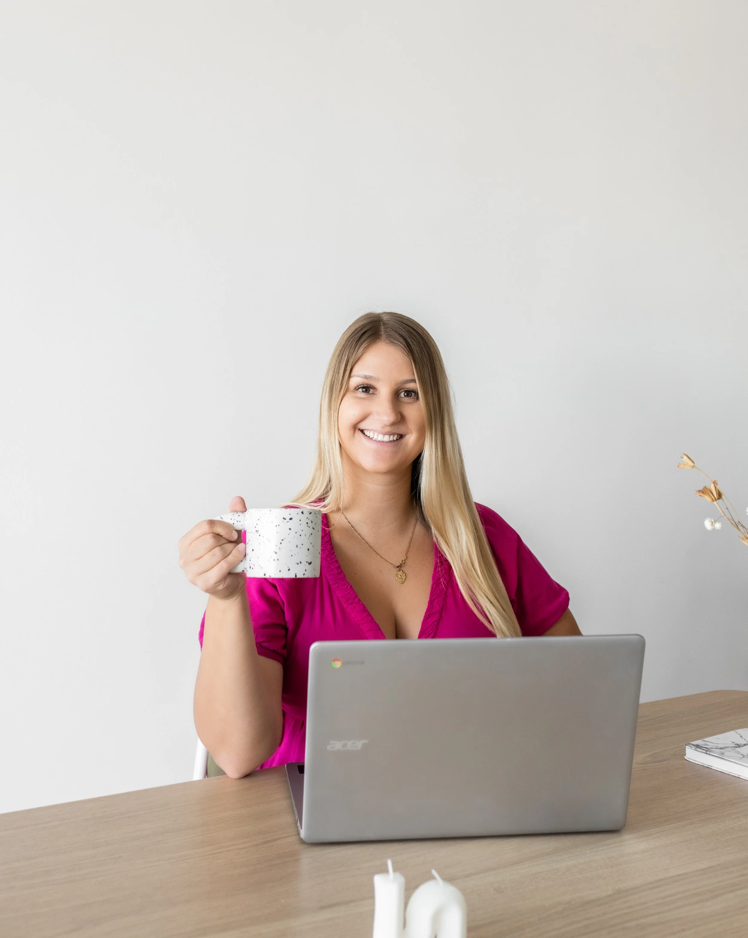 A woman with long blonde hair and a bright pink blouse smiling while holding a white speckled coffee mug near a silver Acer Chromebook laptop on a wooden desk with a white wall background.