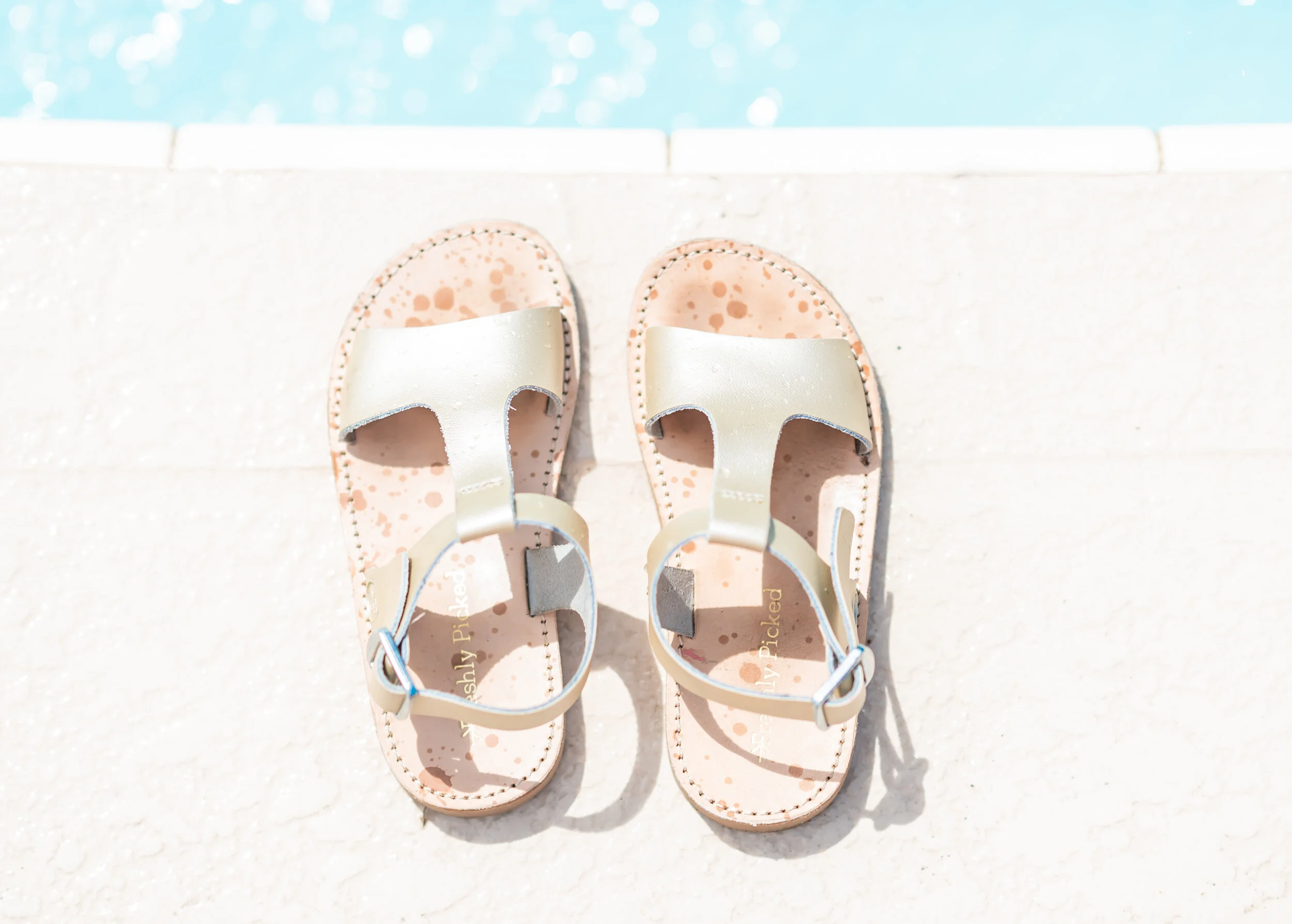 Pair of light-colored sandals with ankle straps on a poolside ledge, with the pool water in the background.