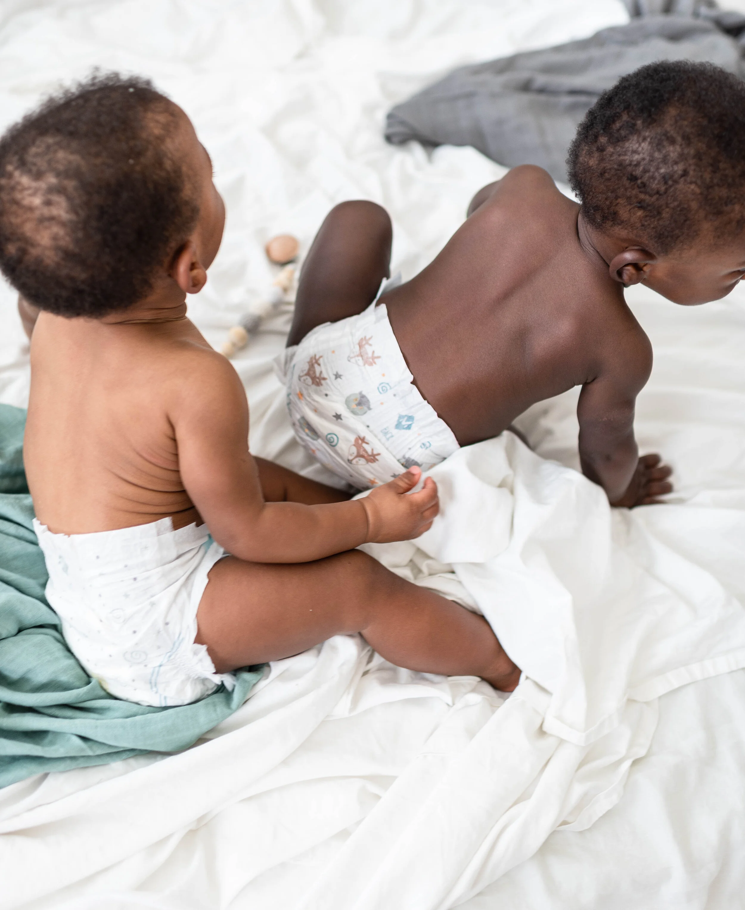 Two young children playing on a bed with white sheets; one is sitting while the other is crawling.