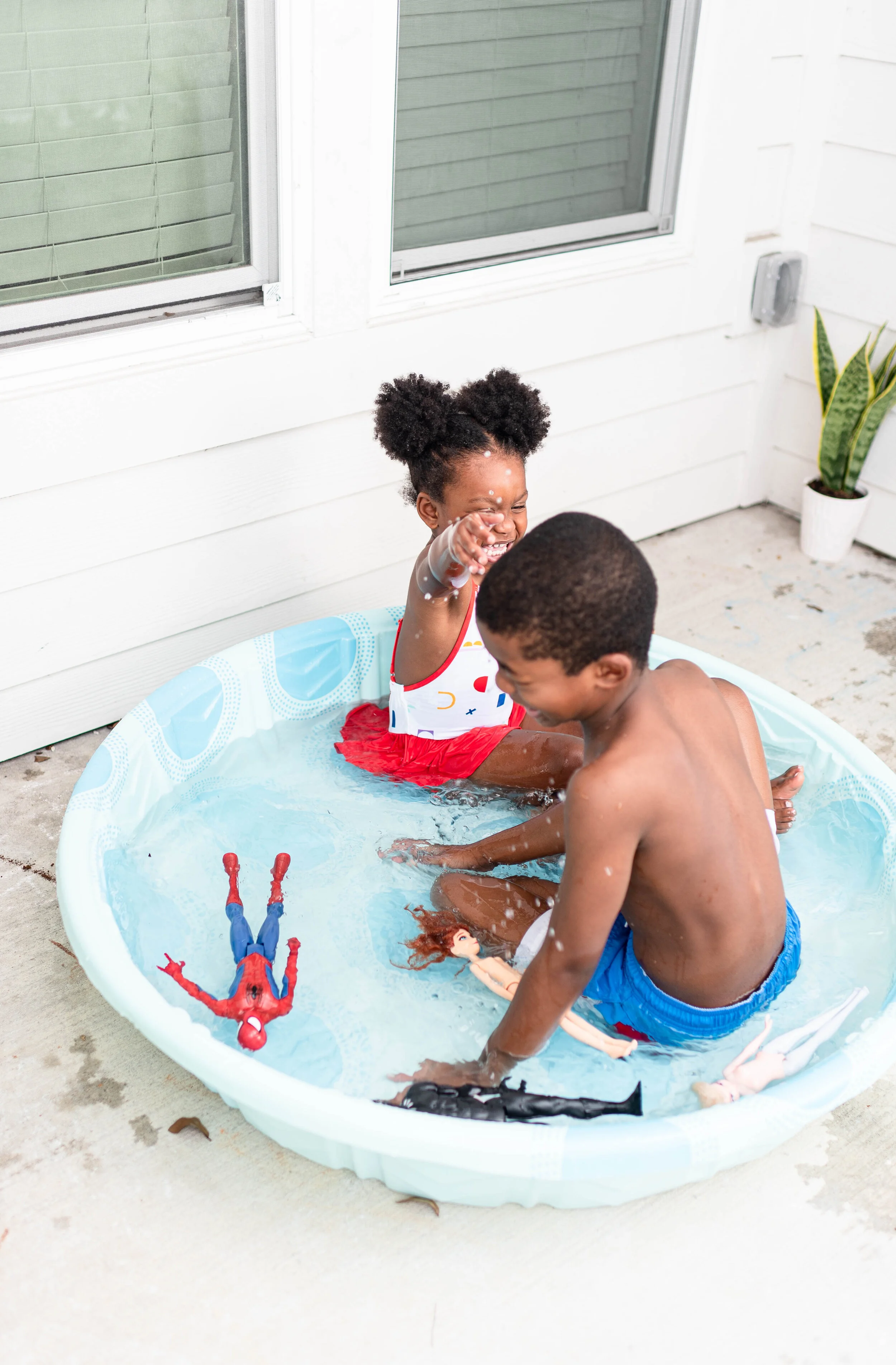 Two children playing in a small inflatable kiddie pool on a porch, laughing and splashing water with toy dolls and a Spider-Man figure floating in the water.