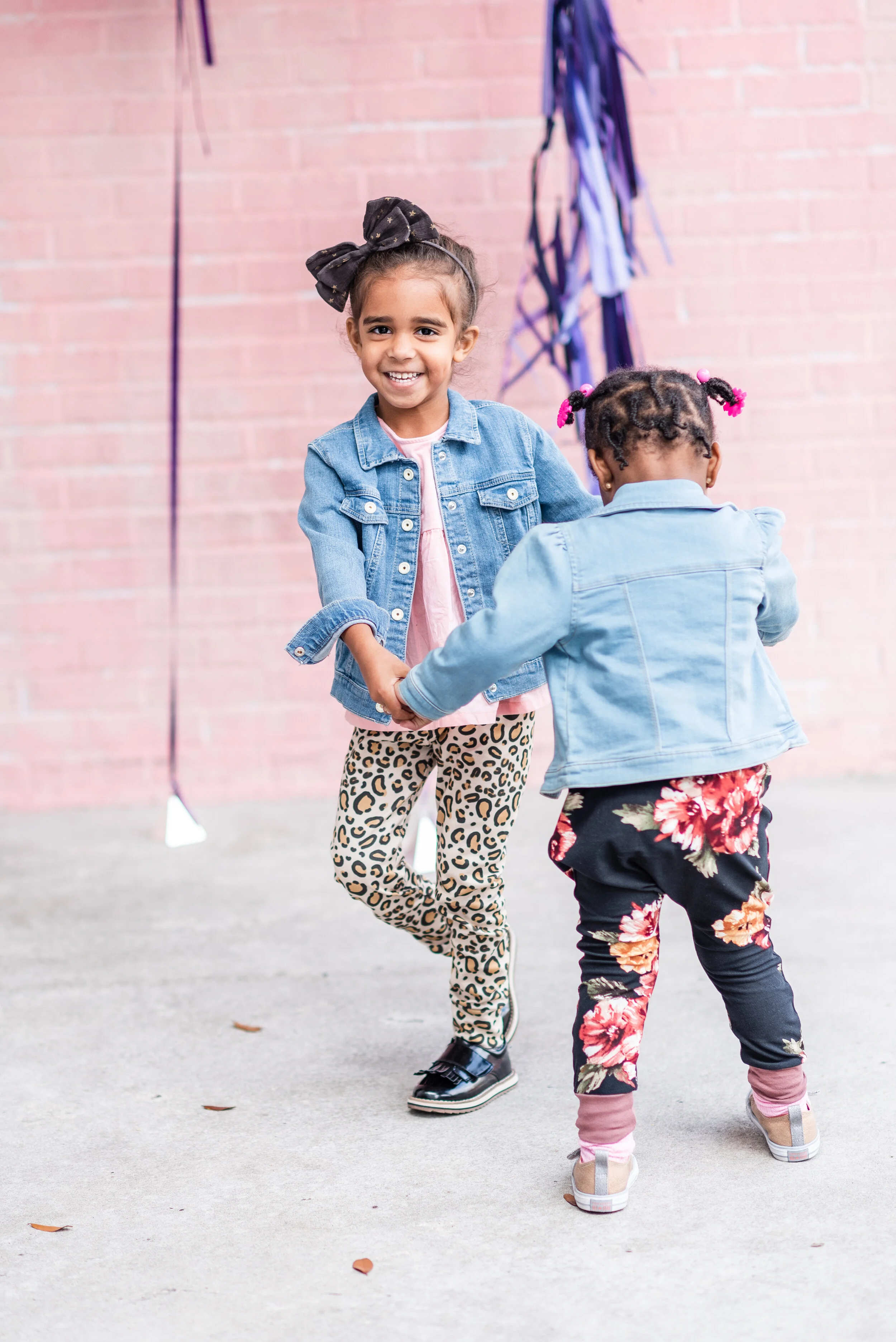 Two young girls are holding hands and dancing or playing together outdoors in front of a pink brick wall. One girl is smiling at the camera while the other has her back turned.