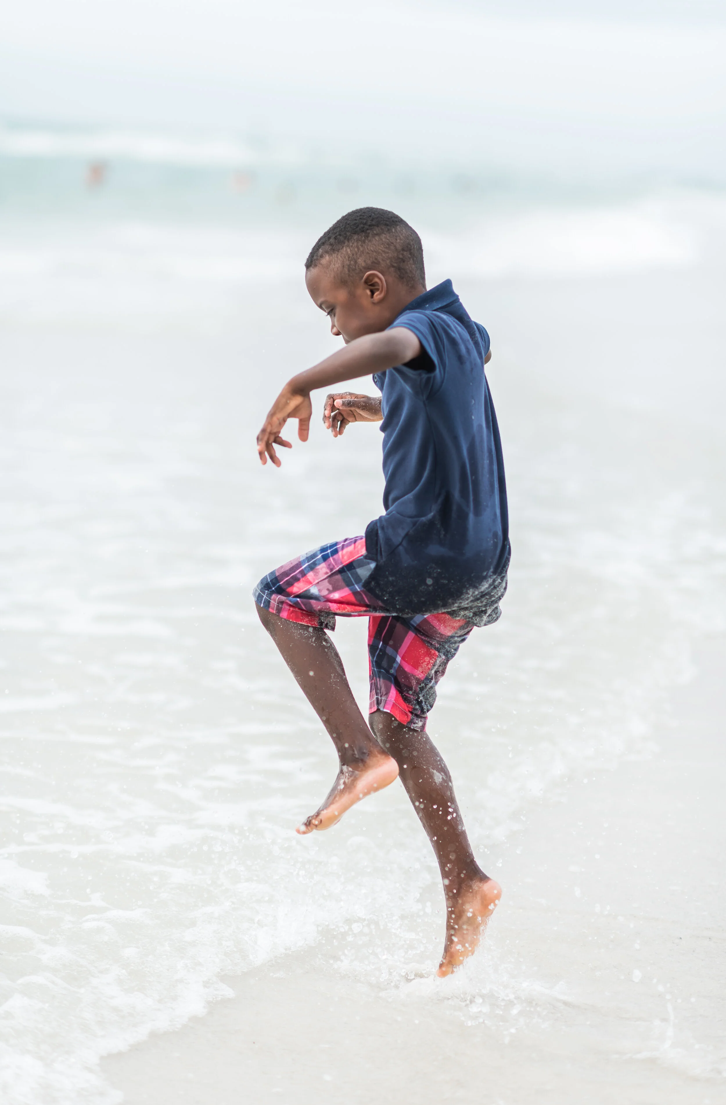 A young boy wearing a navy polo shirt and red plaid shorts playing in shallow ocean water.