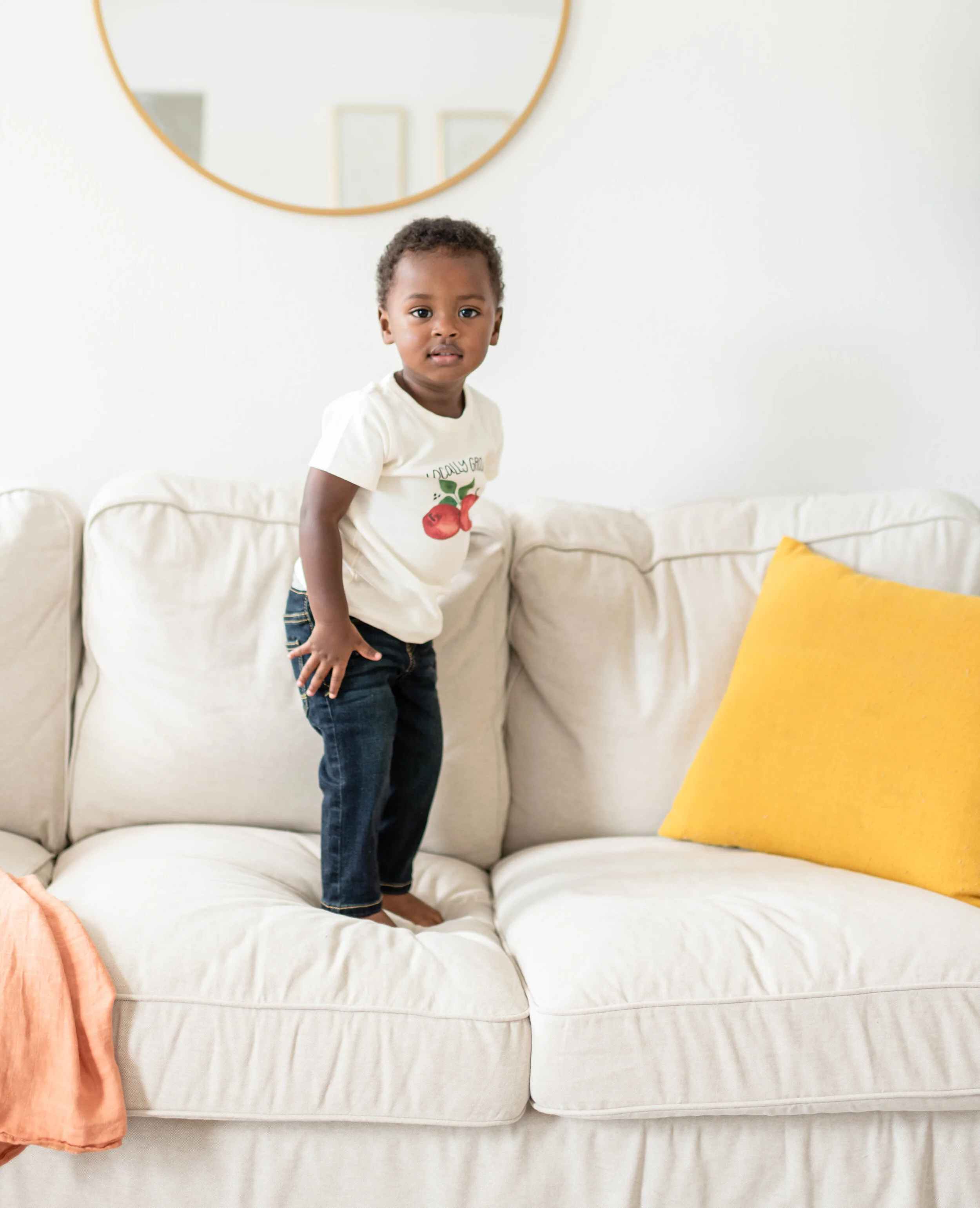 A young child standing on a white couch in a living room with a yellow pillow behind him. The child is wearing a white t-shirt and jeans, and there is a round mirror on the wall behind him.
