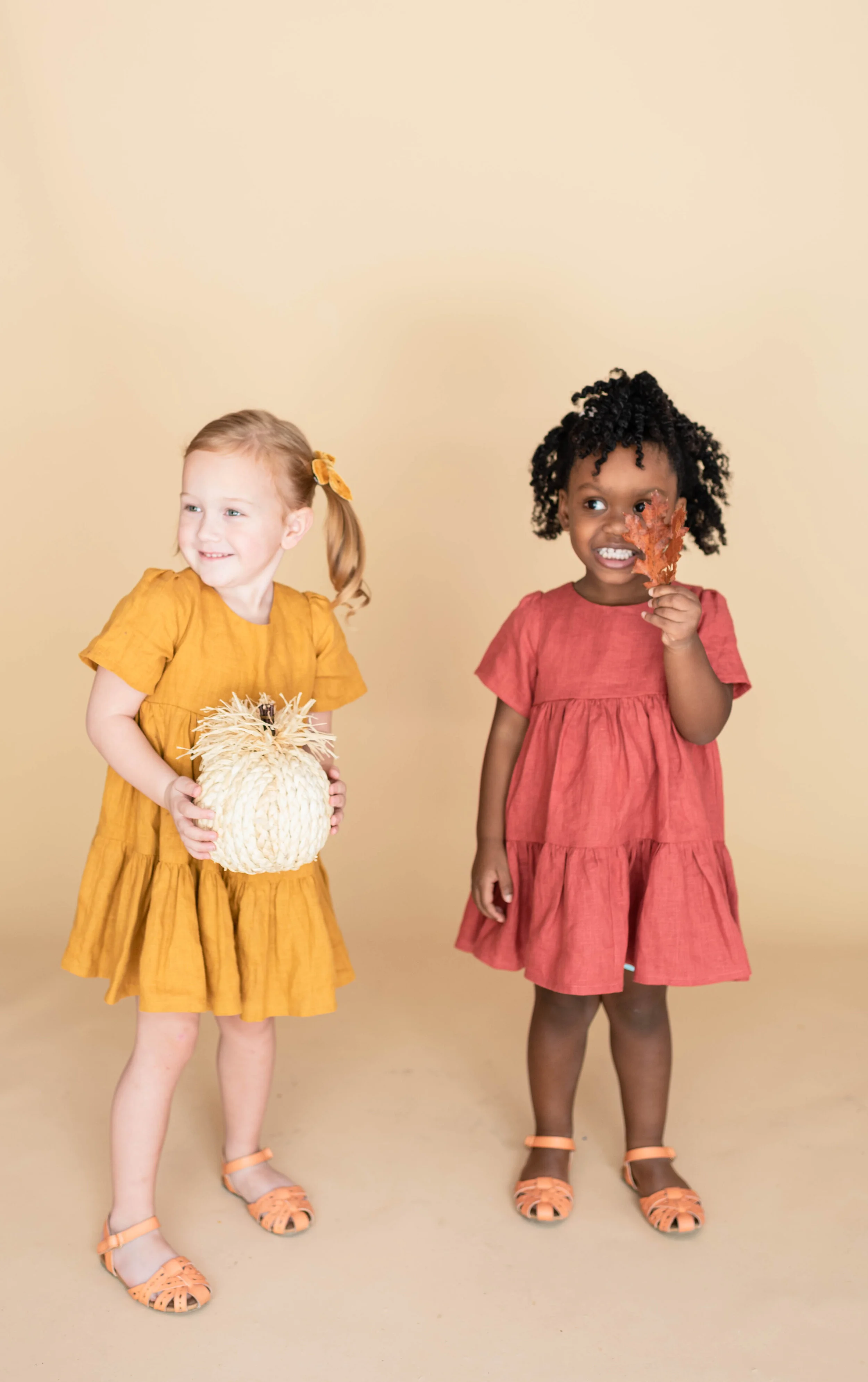Two young girls in orange dresses, one holding a white pumpkin, and the other holding an autumn leaf.