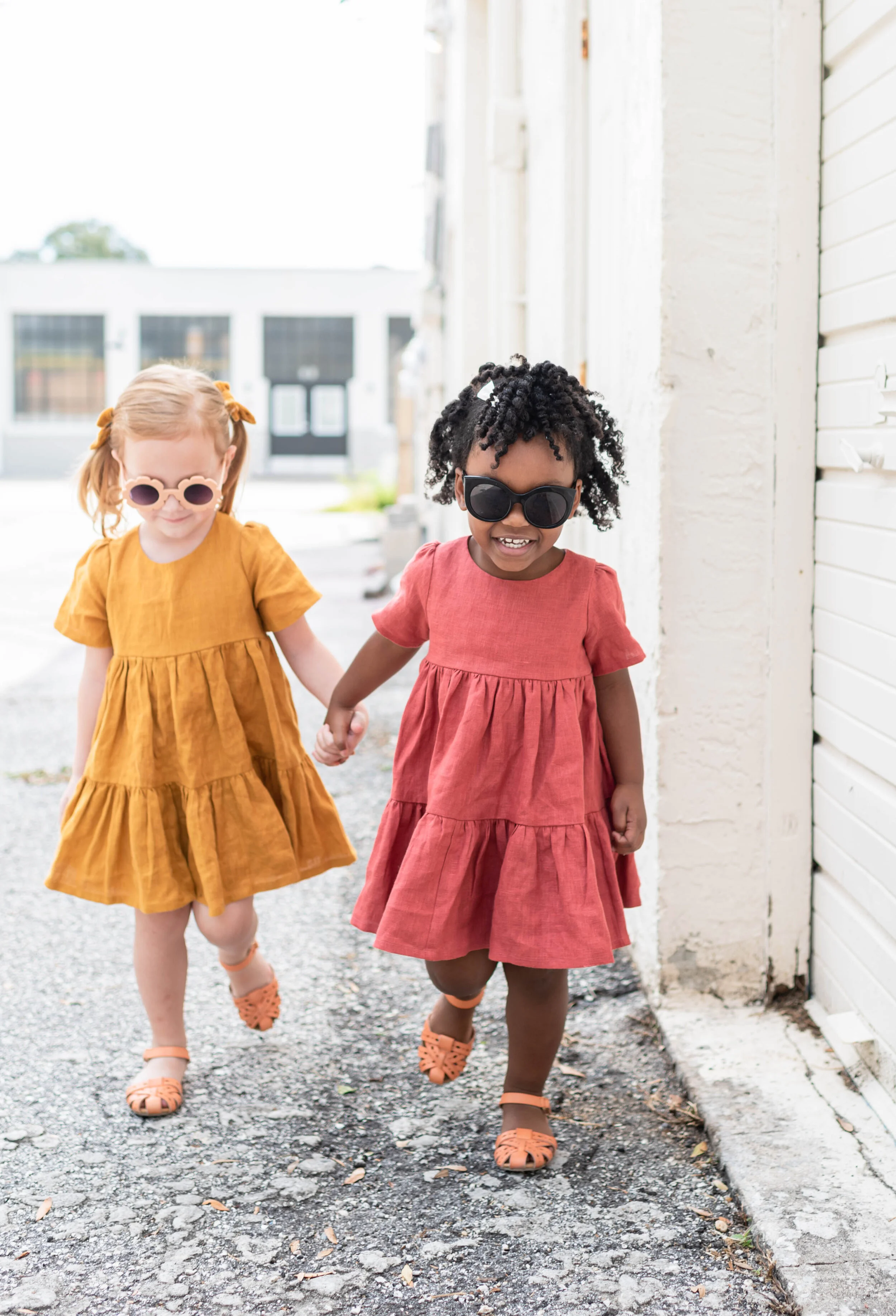 Two young girls wearing colorful dresses and sunglasses holding hands and walking outside on a sunny day.