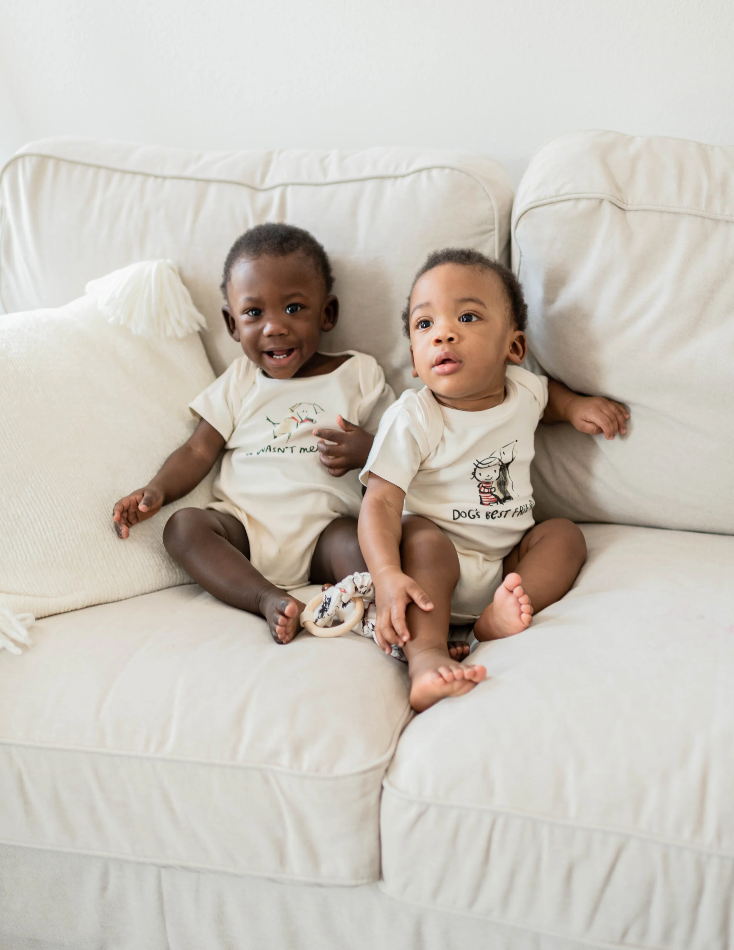 Two young children sitting on a white sofa, wearing matching cream-colored shirts, one smiling and the other looking surprised or curious.