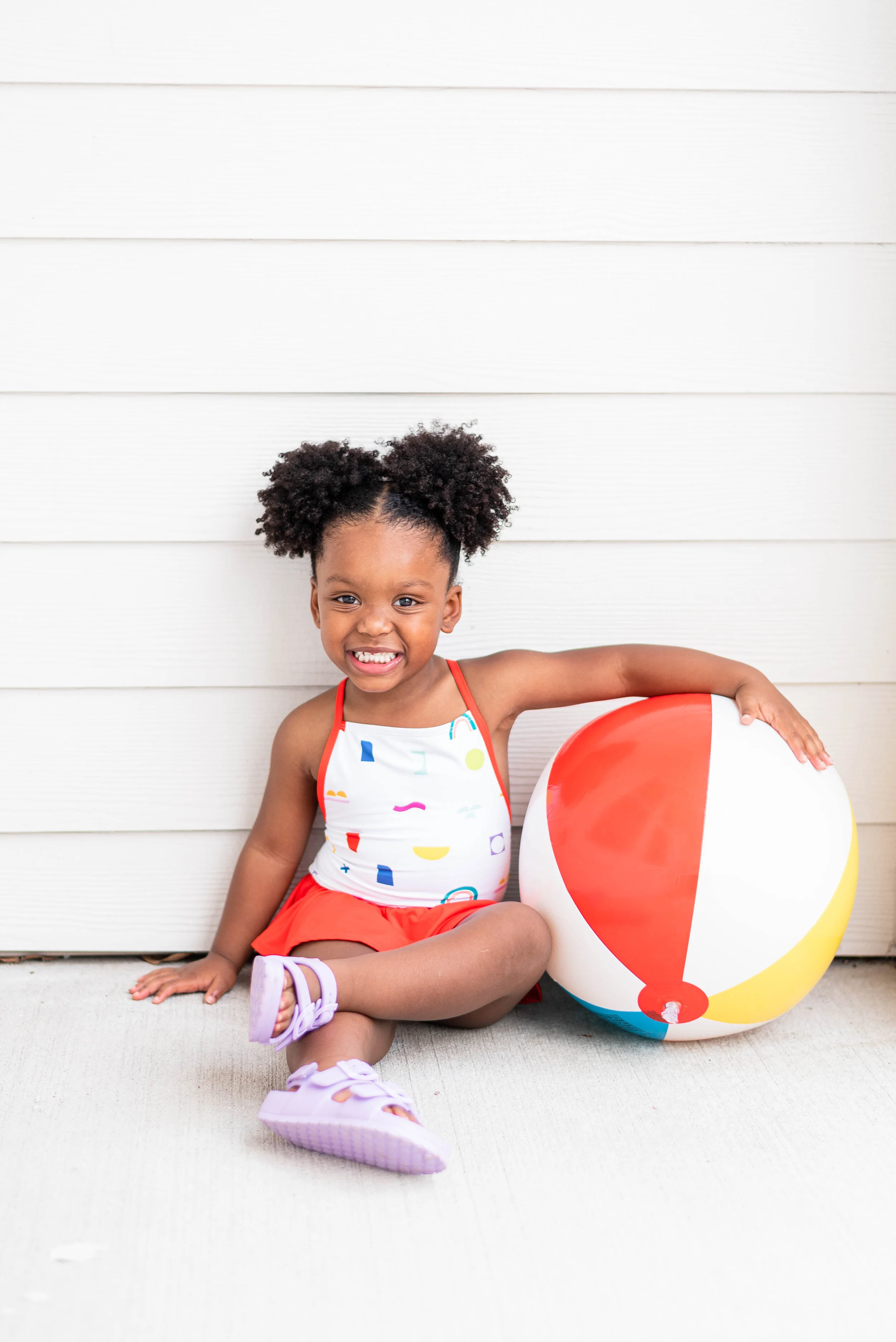 A young girl with curly hair smiling, sitting on the ground with her legs crossed next to a colorful beach ball, in front of a white wooden wall.