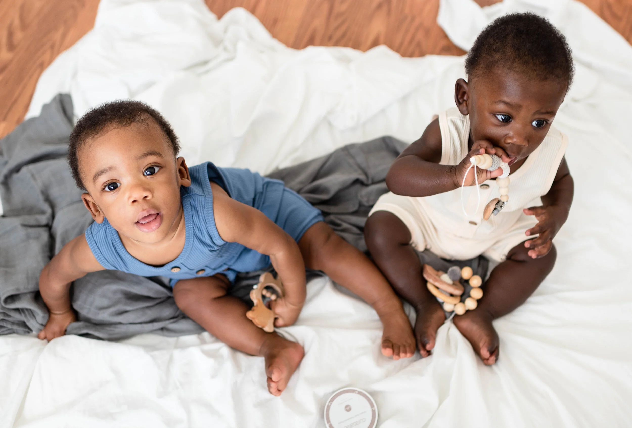 Two young children sit on a bed covered with a white sheet, with gray and white blankets, playing with teething toys.