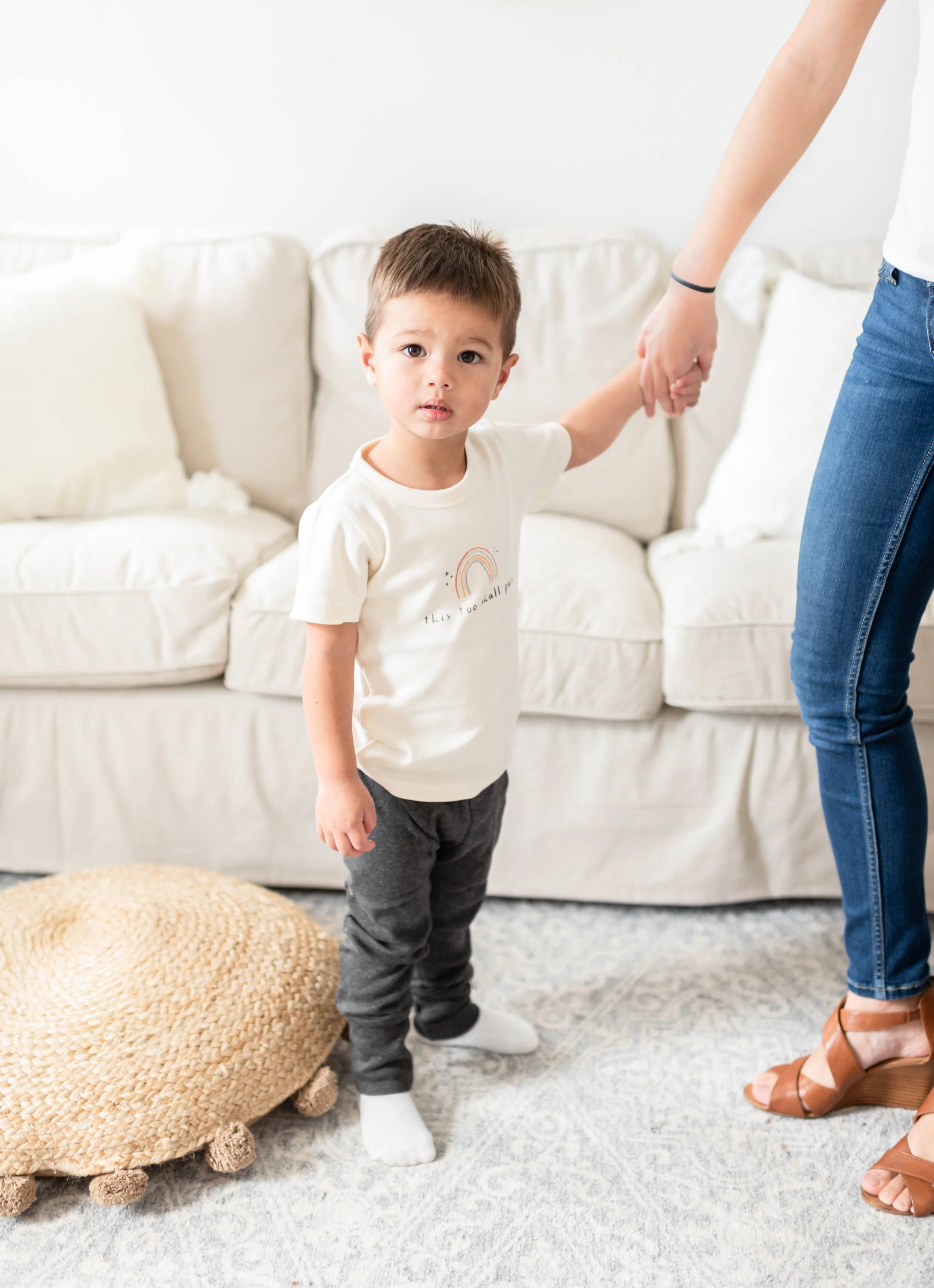 A young boy stands on a patterned rug, holding hands with an adult. The boy wears a white T-shirt with a rainbow graphic and the words "this too shall pass," gray pants, and white socks. The adult wears blue jeans and tan sandals. There is a beige wo