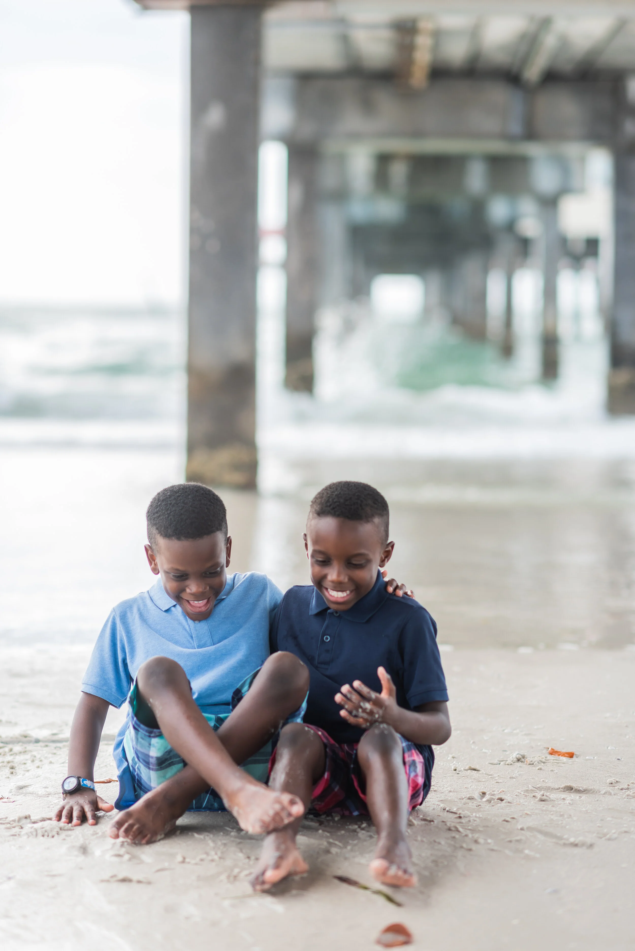 Two young boys in blue shirts and shorts playing and laughing on the sandy beach under a pier.