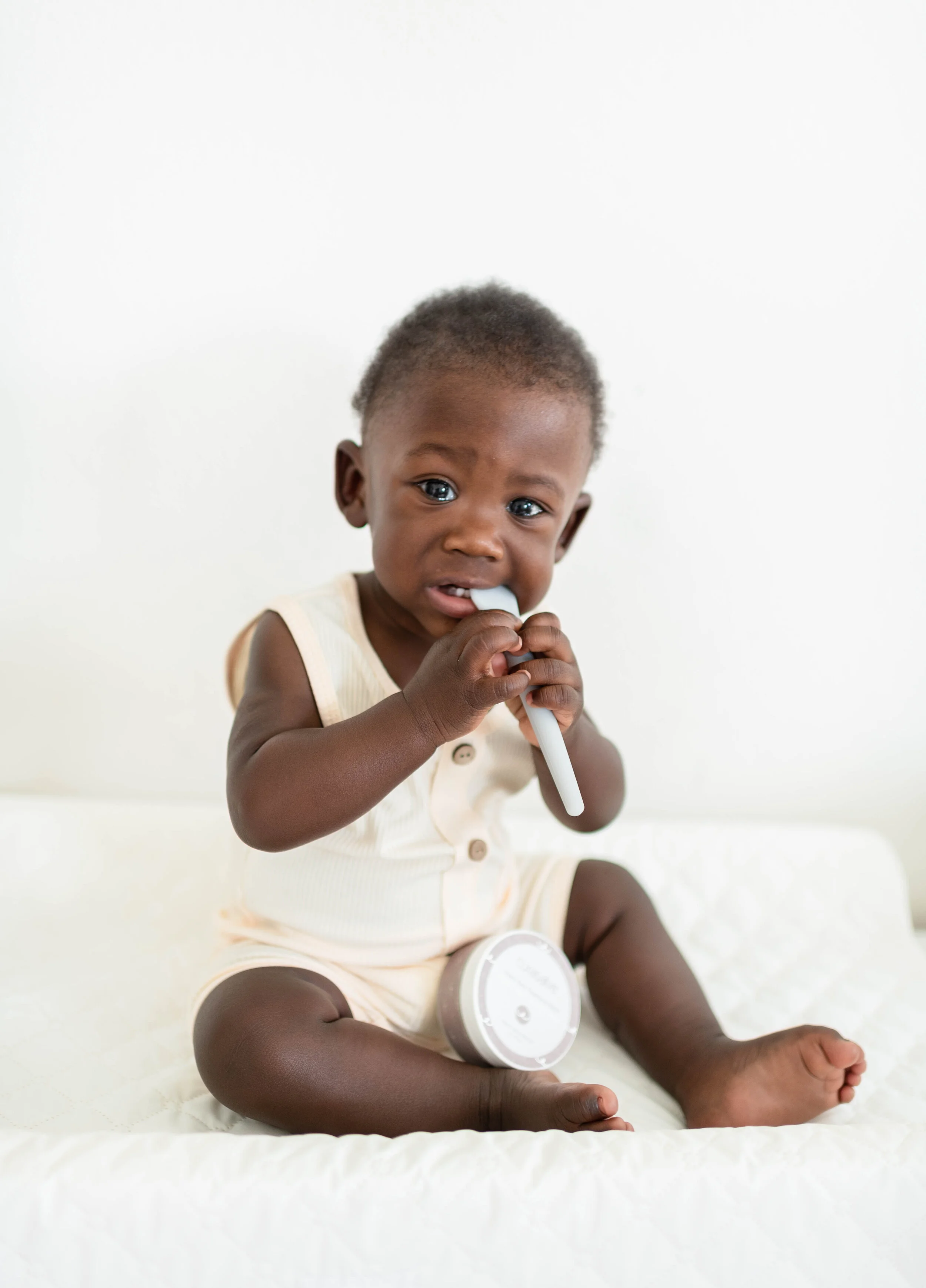 A young African American toddler sitting on a white surface, holding and chewing a toothbrush, with a container of baby cream or lotion nearby. The background is plain white.