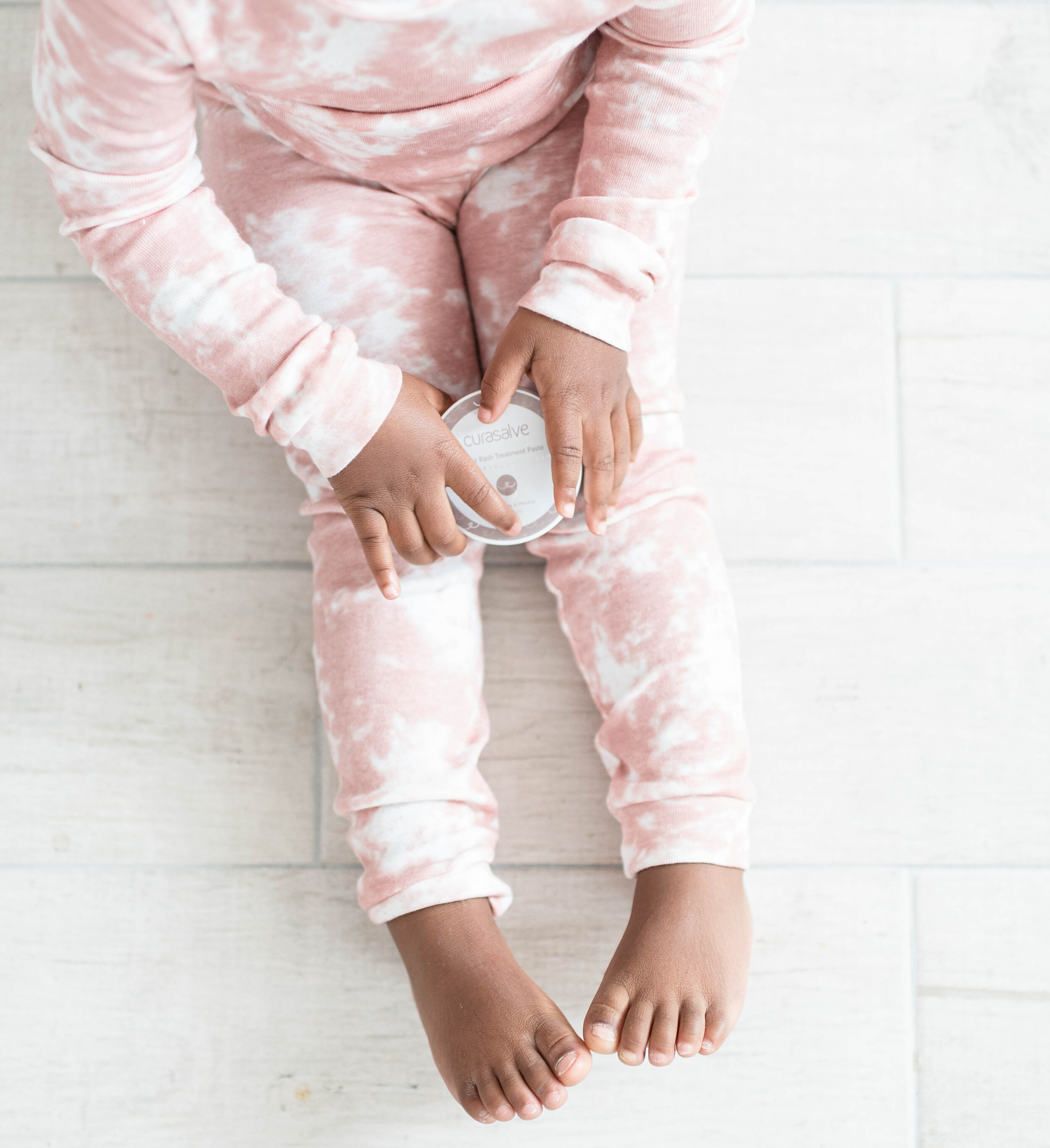 A child wearing pink and white tie-dye pajamas is sitting on a light-colored wooden floor, holding a container of Curasalve rash treatment paste.