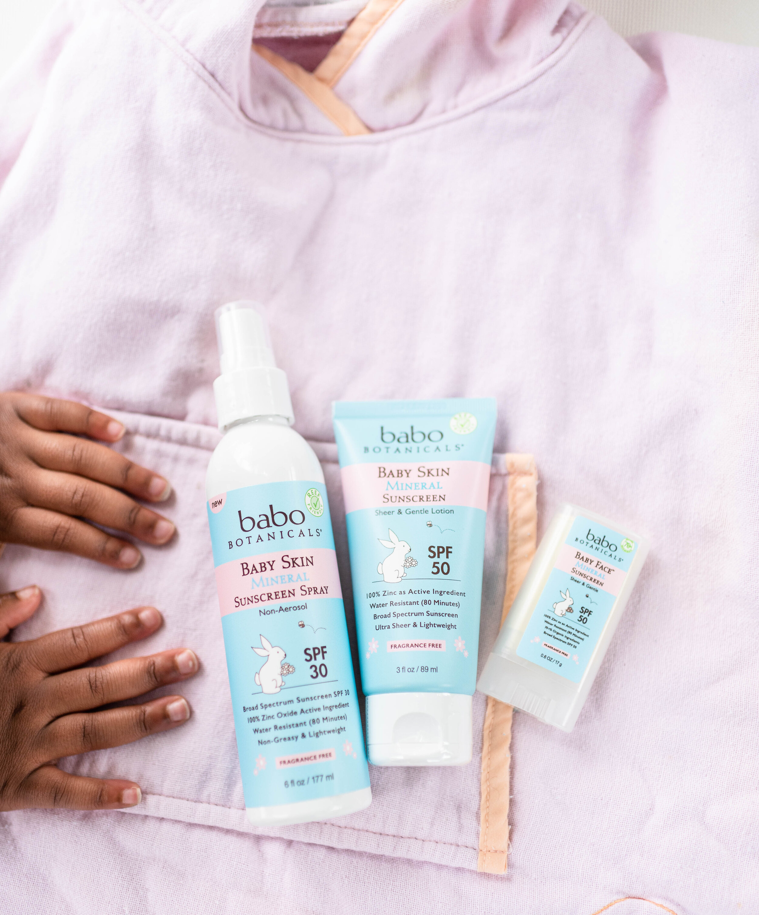 Three skincare products from Babo Botanicals for babies with sun protection, including sunscreen spray, lotion, and stick, placed on a pink fabric surface. A child's hand is reaching toward the products.