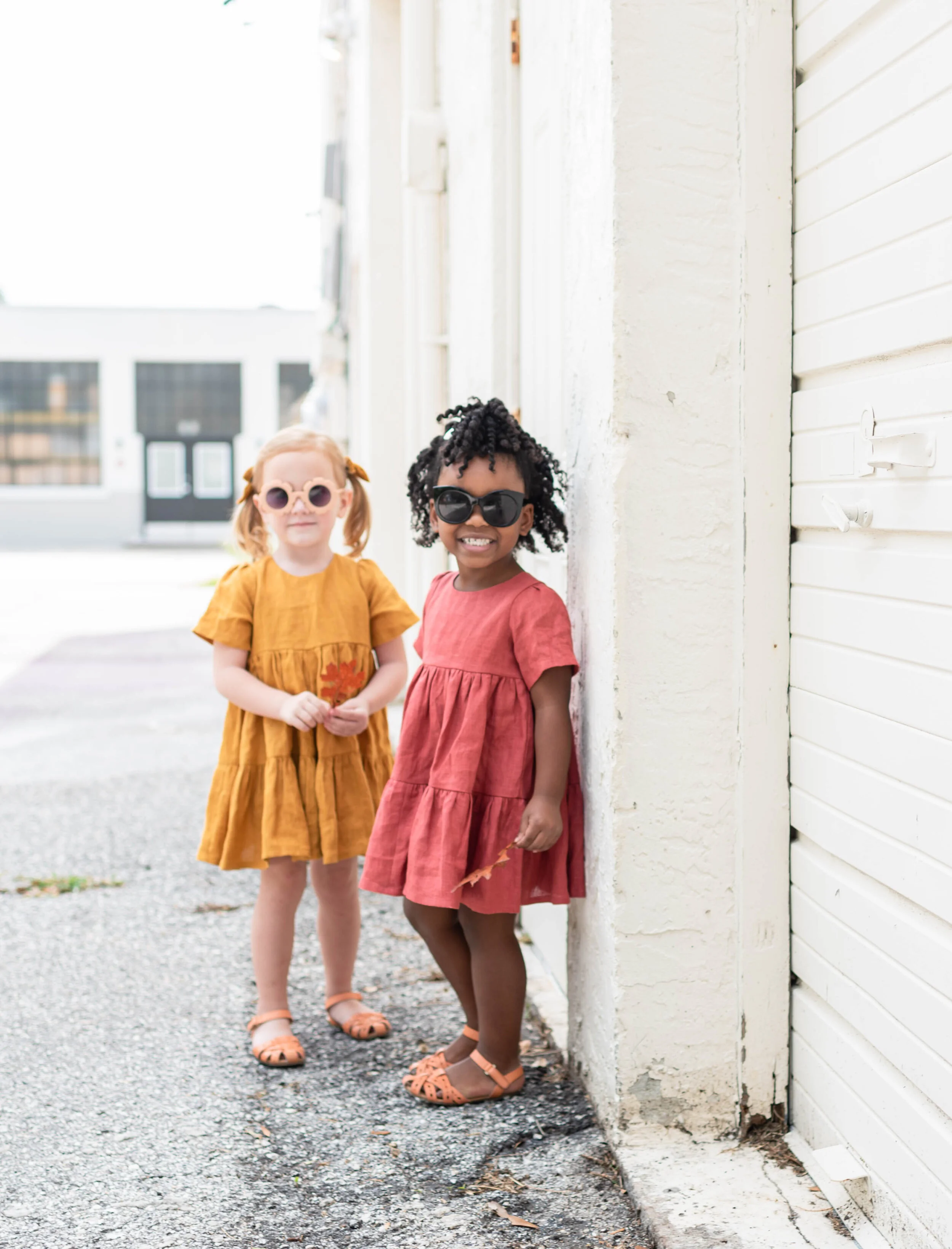 Two young girls wearing colorful dresses and sunglasses, standing on a sidewalk next to a white building with white siding, smiling at the camera.