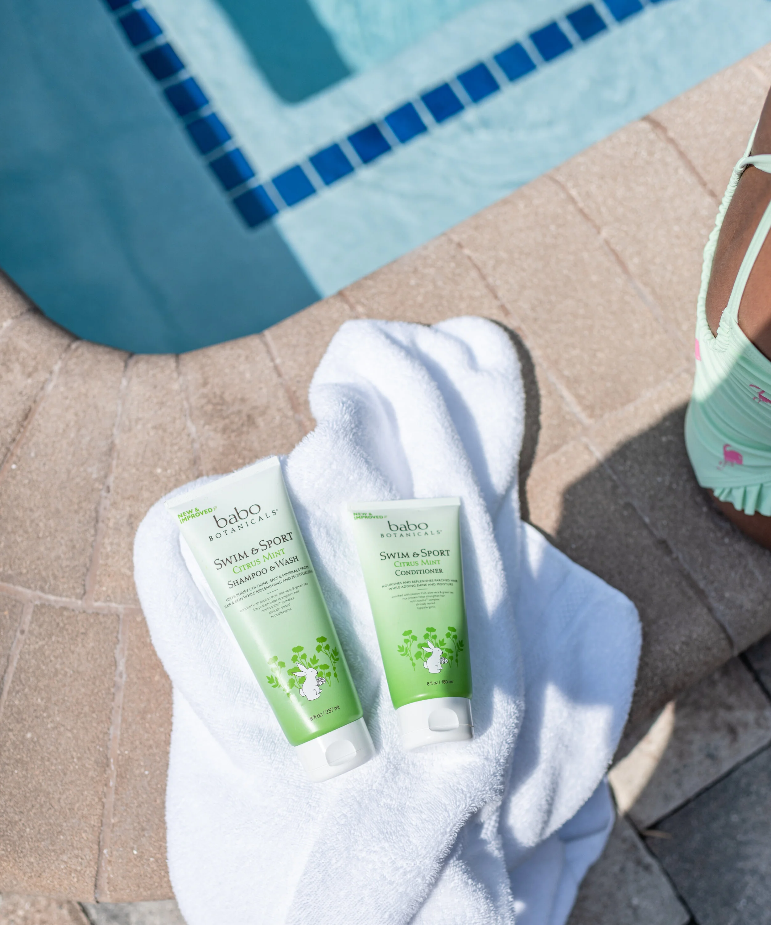 Two tubes of Babo Botanicals Swim & Sport Citrus Mint shampoo and conditioner resting on a white towel near a swimming pool, with part of a woman's foot wearing a beach sandal visible.