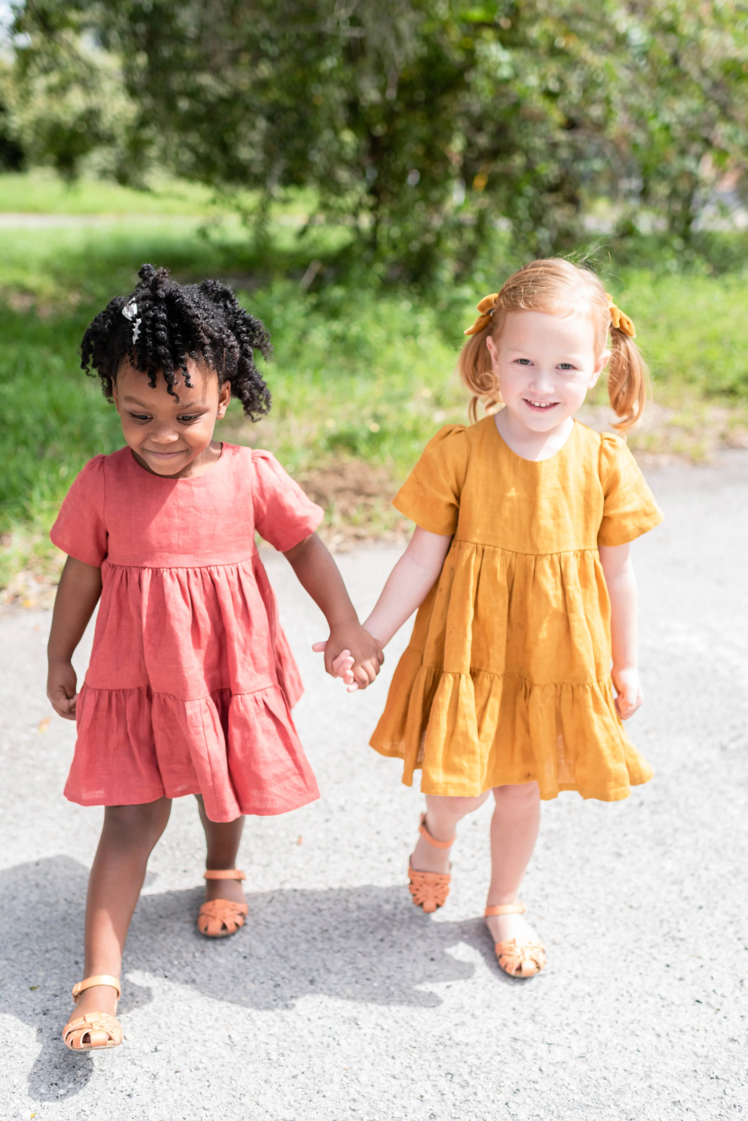Two young girls holding hands and walking outdoors on a bright day, with greenery and a tree in the background. One girl has curly black hair and is wearing a pink dress, while the other girl has red hair tied with yellow ribbons and is wearing a yel