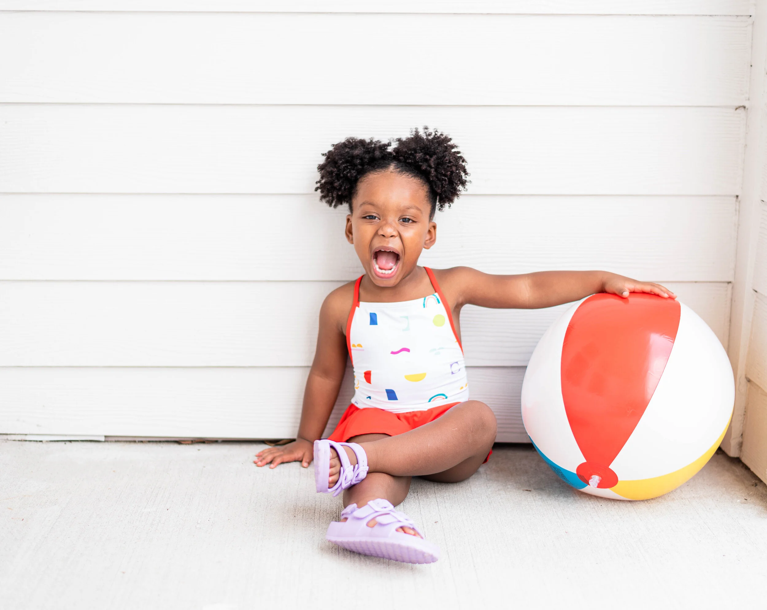 A young girl with curly hair sitting on the floor next to a beach ball, wearing colorful clothing, and making a playful expression.
