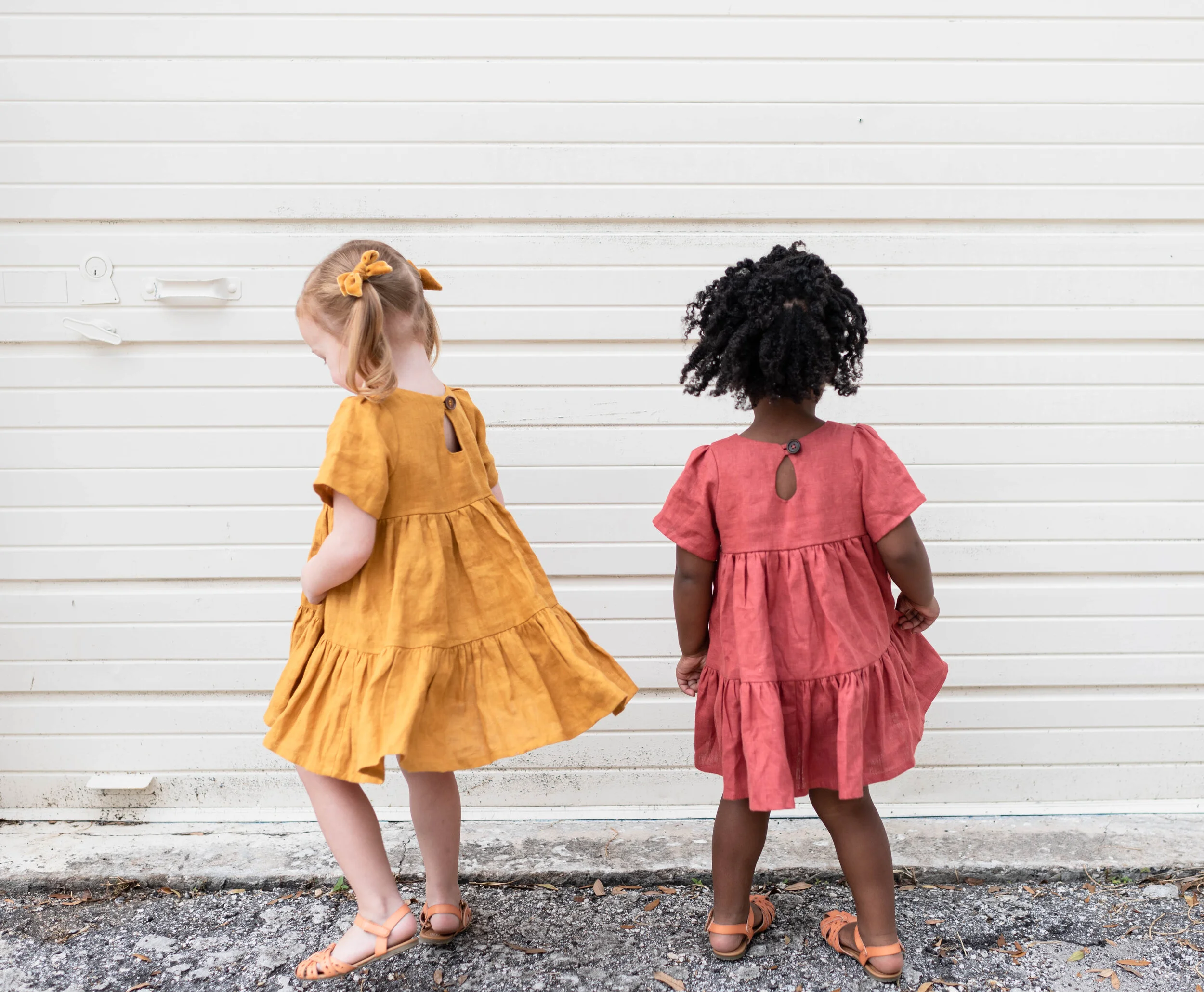 Two young girls standing with their backs to the camera, wearing colorful dresses and sandals, in front of a white horizontal siding wall.