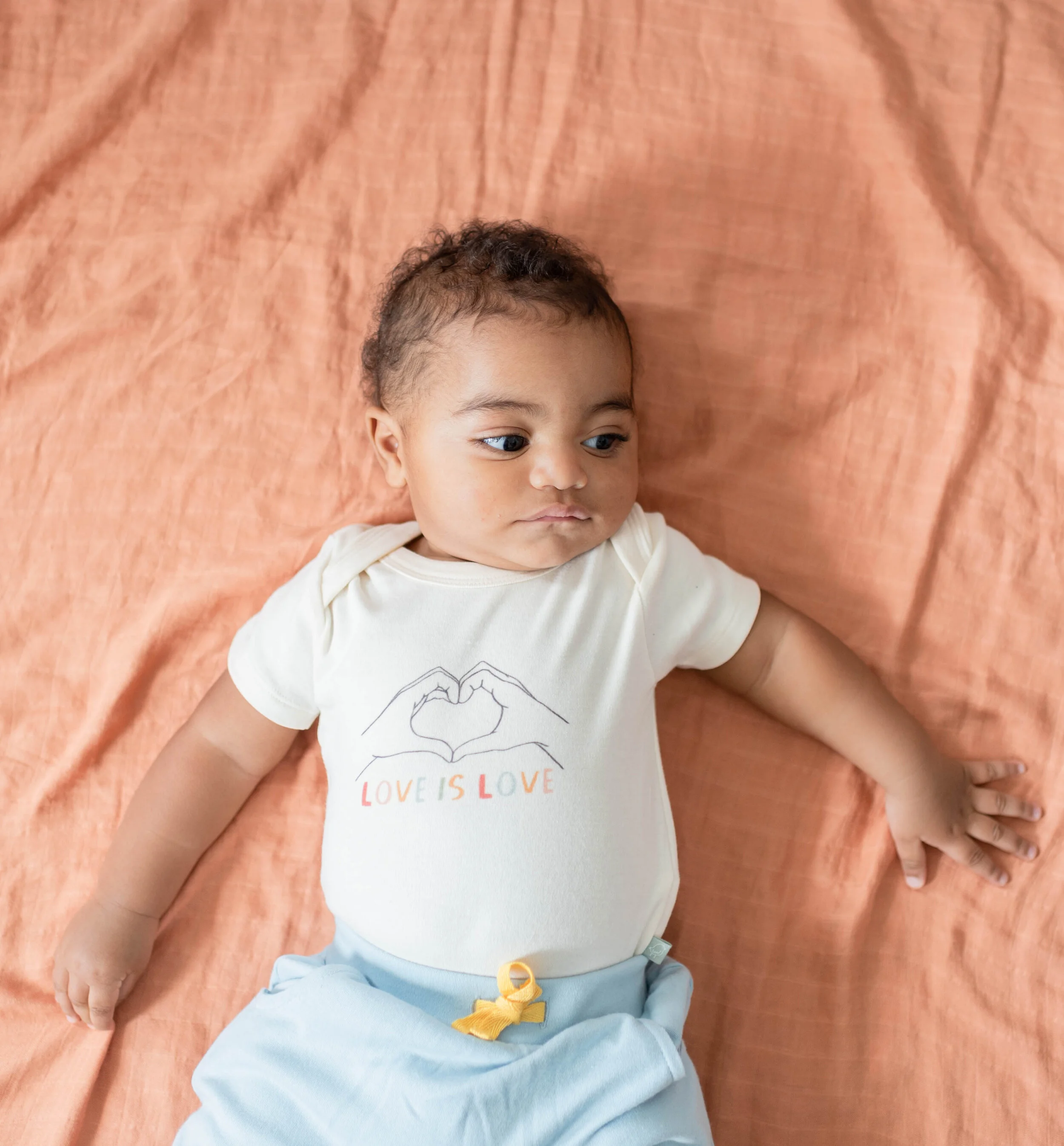 A baby lying on a peach-colored blanket, wearing a white T-shirt with a drawing of hands forming a heart and the words "LOVE IS LOVE" underneath, and light blue pants with a yellow ribbon tied around the waistband.
