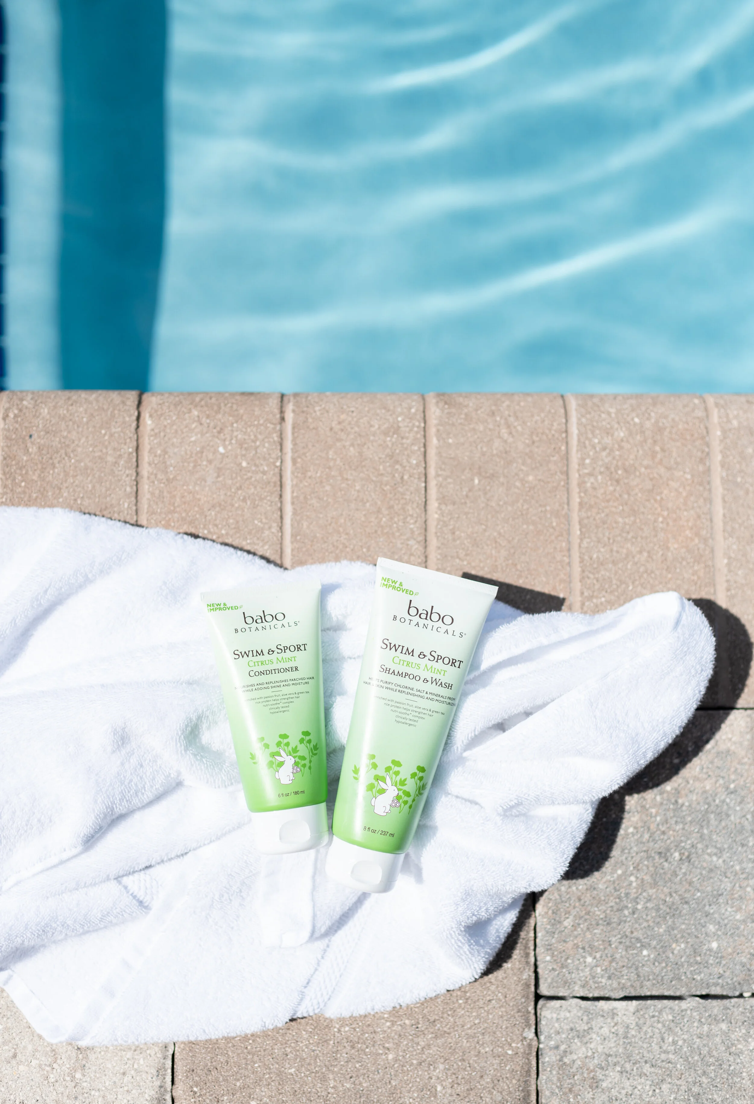 Two tubes of Babo Botanicals Swim & Sport Citrus Mint shampoo and conditioner on a white towel next to a swimming pool.