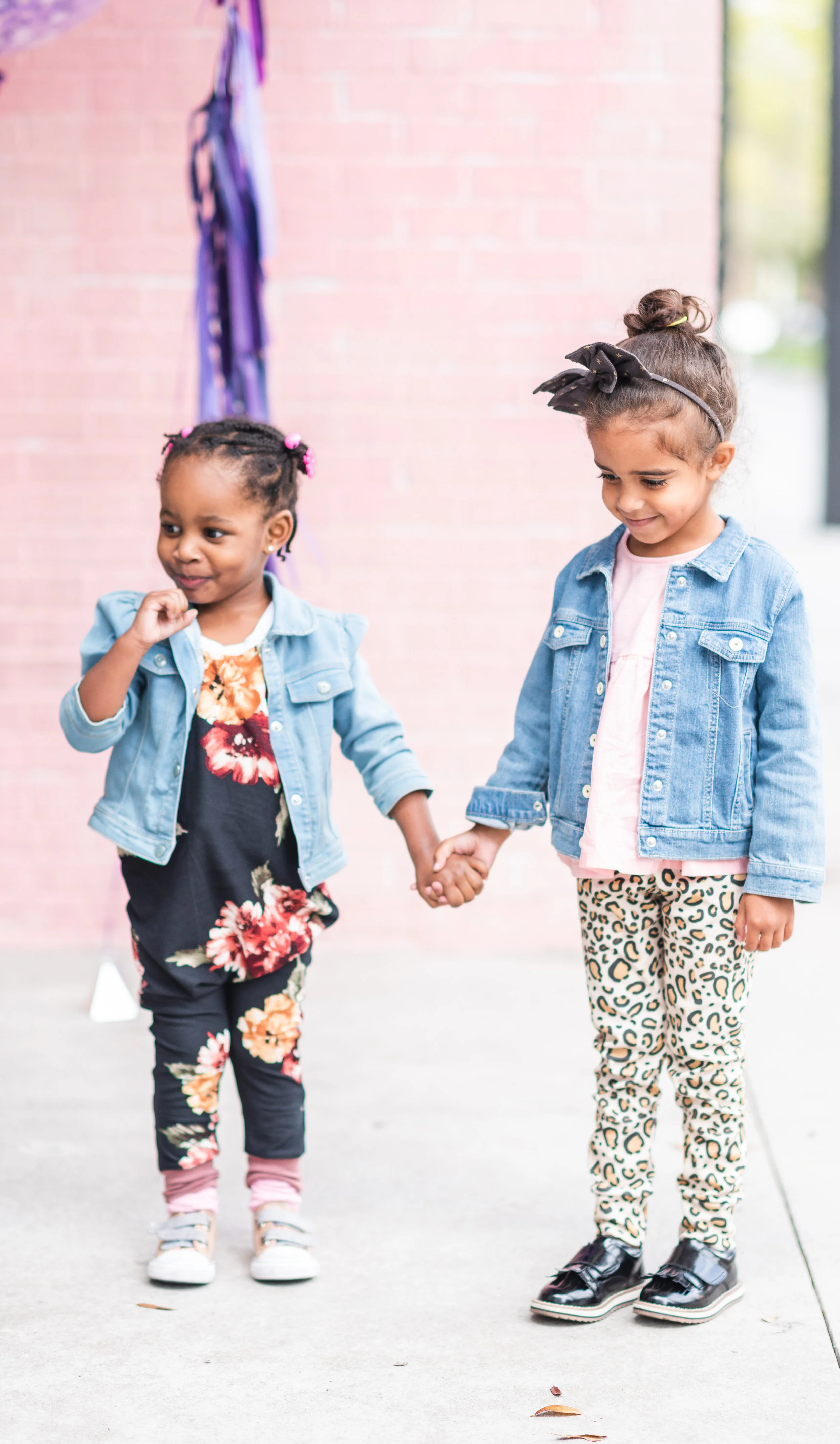 Two young girls holding hands and smiling outdoors, wearing denim jackets and patterned leggings.