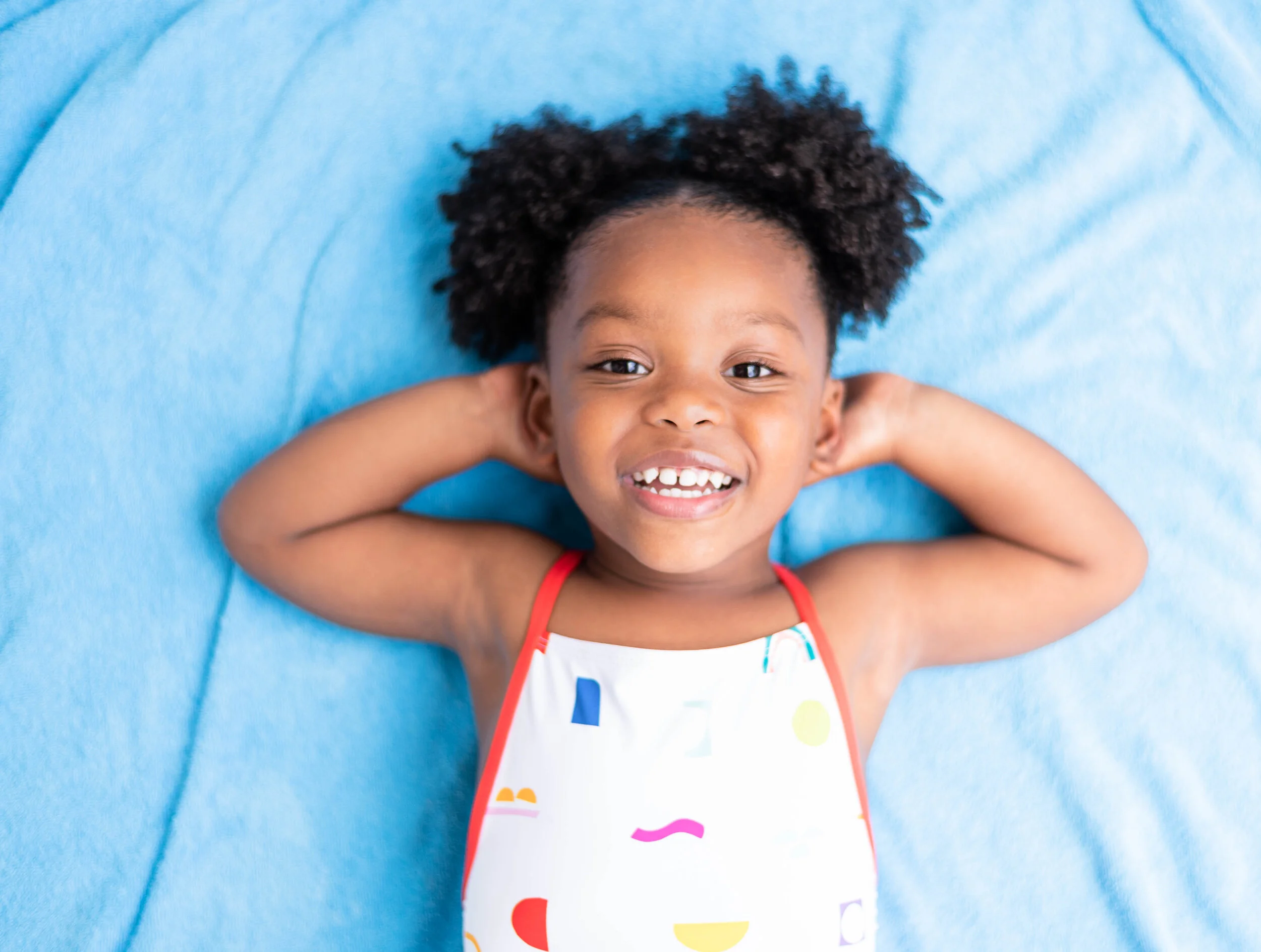 A young girl lying on her back on a blue blanket, smiling, with her hands behind her head, wearing a colorful sleeveless top.