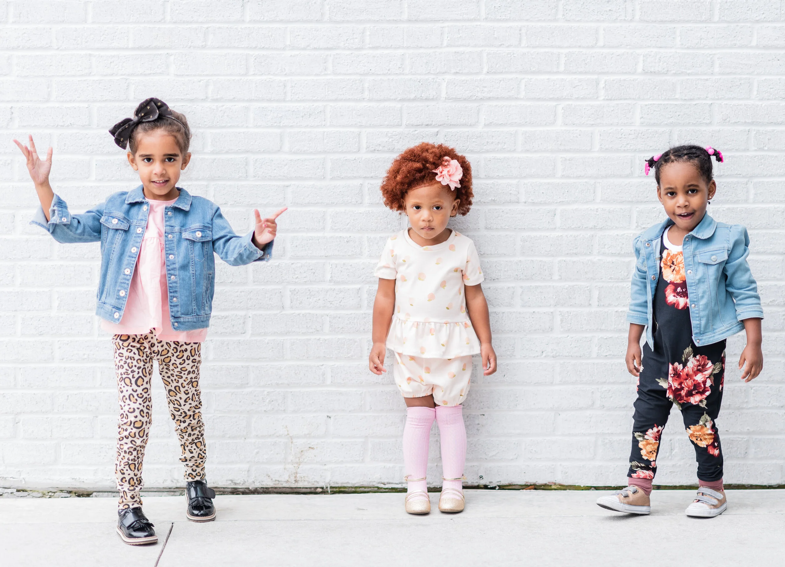 Three young girls standing in front of a white brick wall, wearing casual outfits and denim jackets, with varied facial expressions.