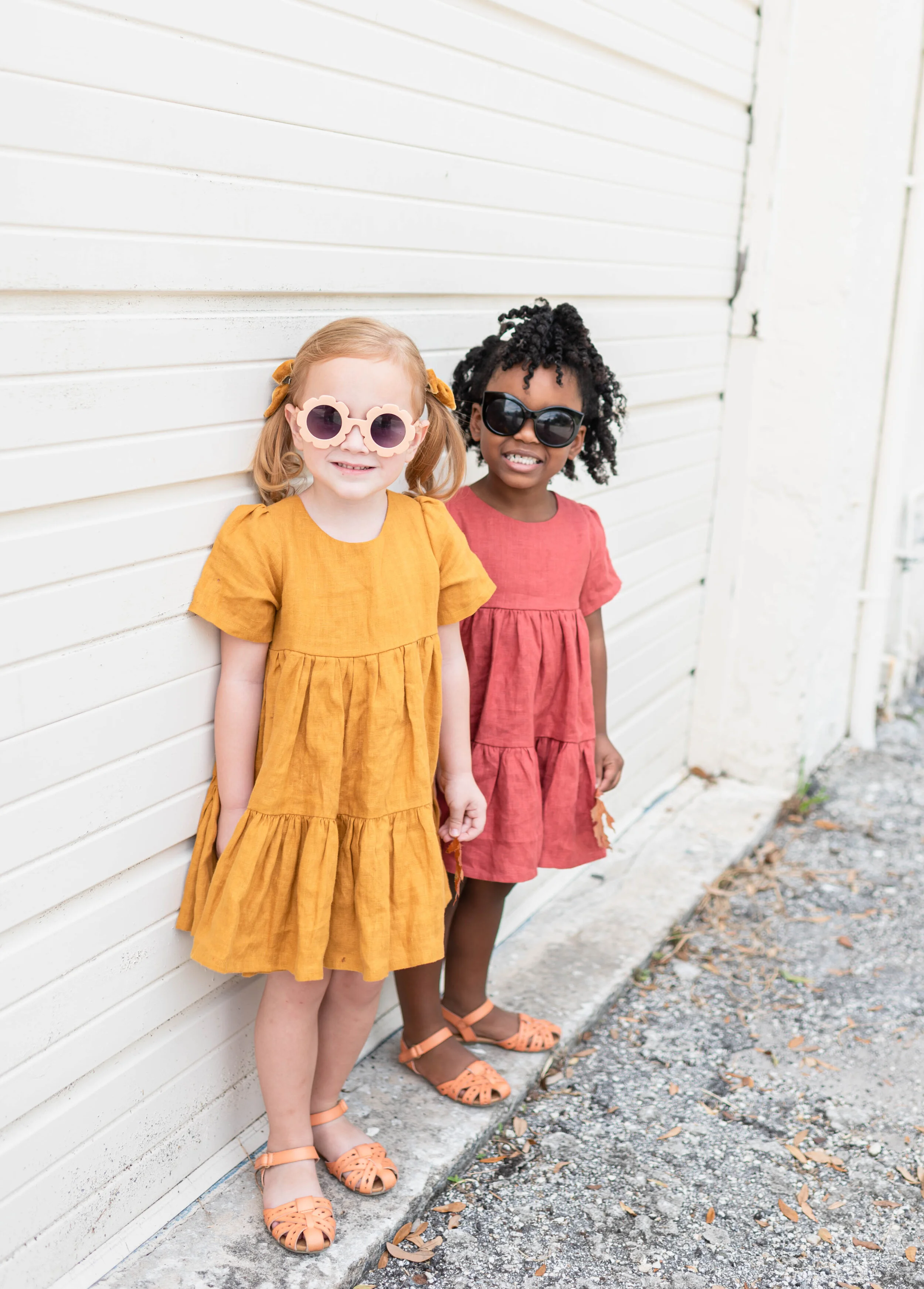 Two young girls wearing colorful dresses and sunglasses standing against a white wooden wall outdoors.
