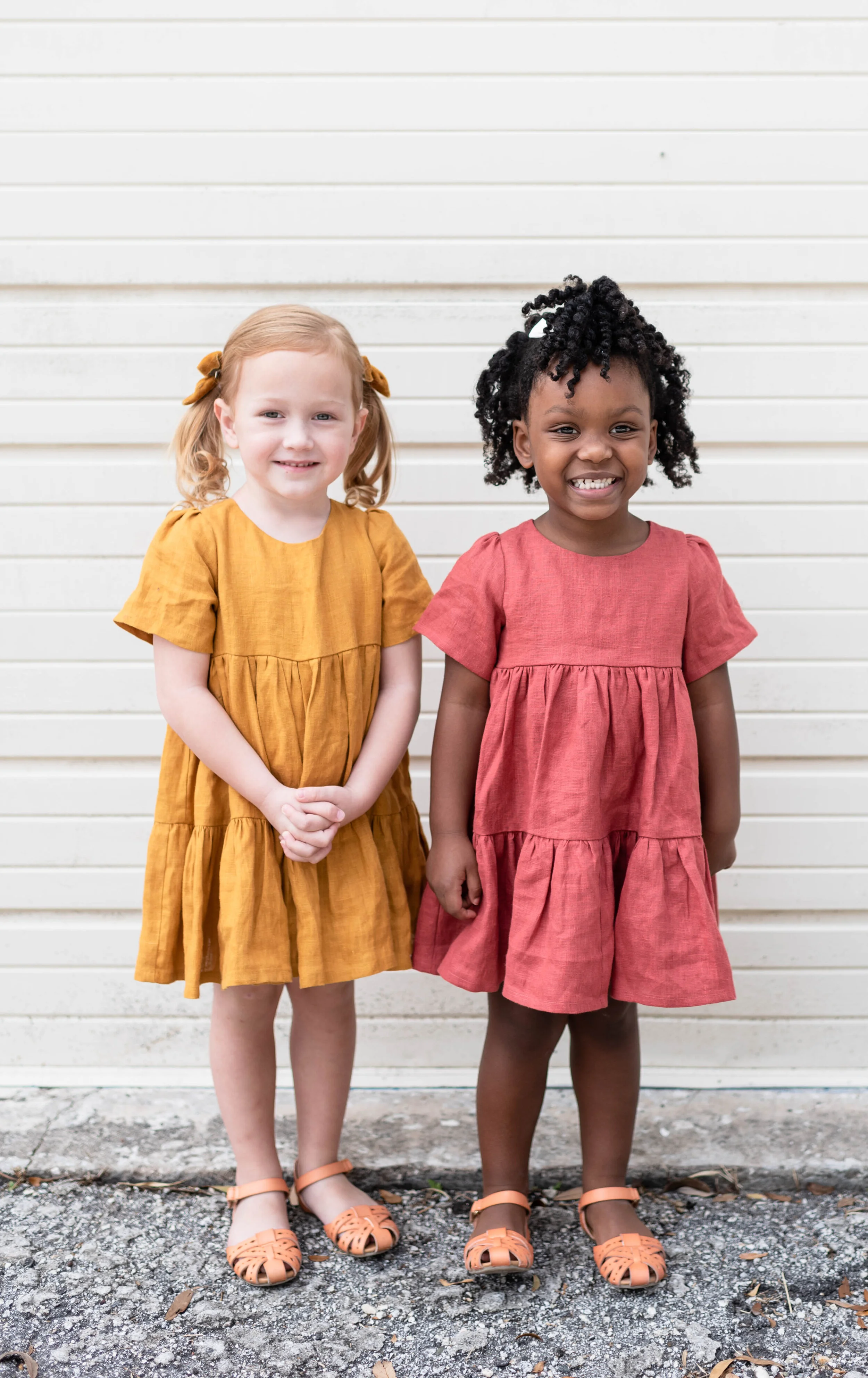 Two young girls standing outdoors in front of a white horizontal wall, smiling at the camera. One girl has blonde hair, and the other has black curly hair. Both are wearing colorful dresses and sandals.