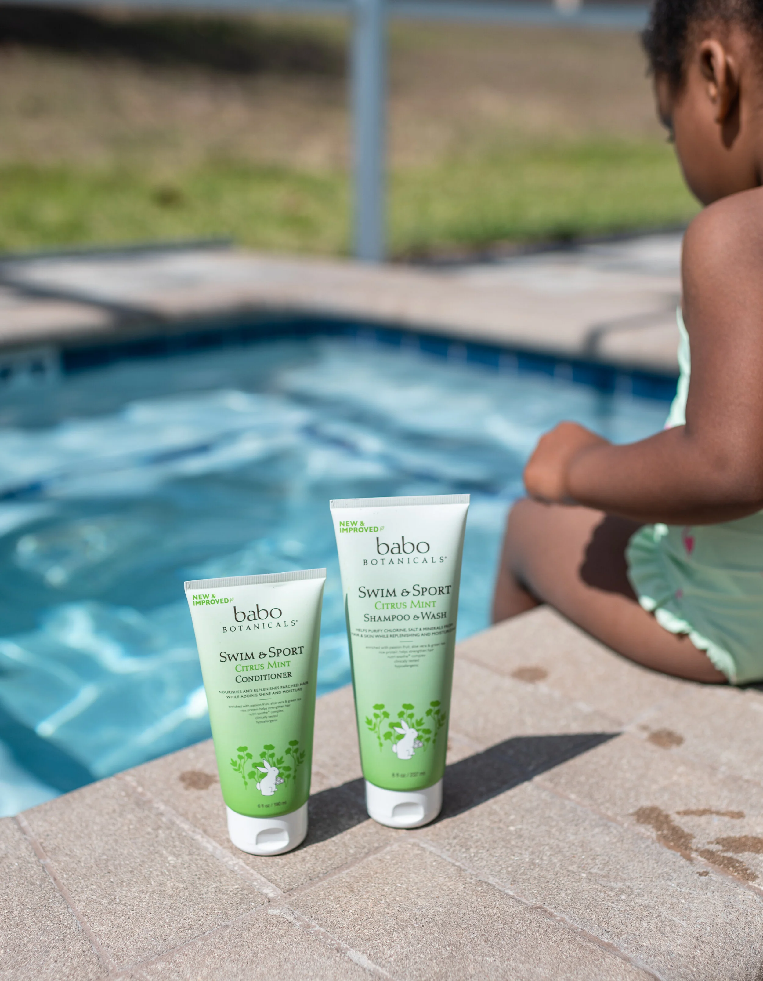 Two tubes of Babo Botanicals Swim & Sport Citrus Mint shampoo and conditioner on poolside with a child sitting by the pool.