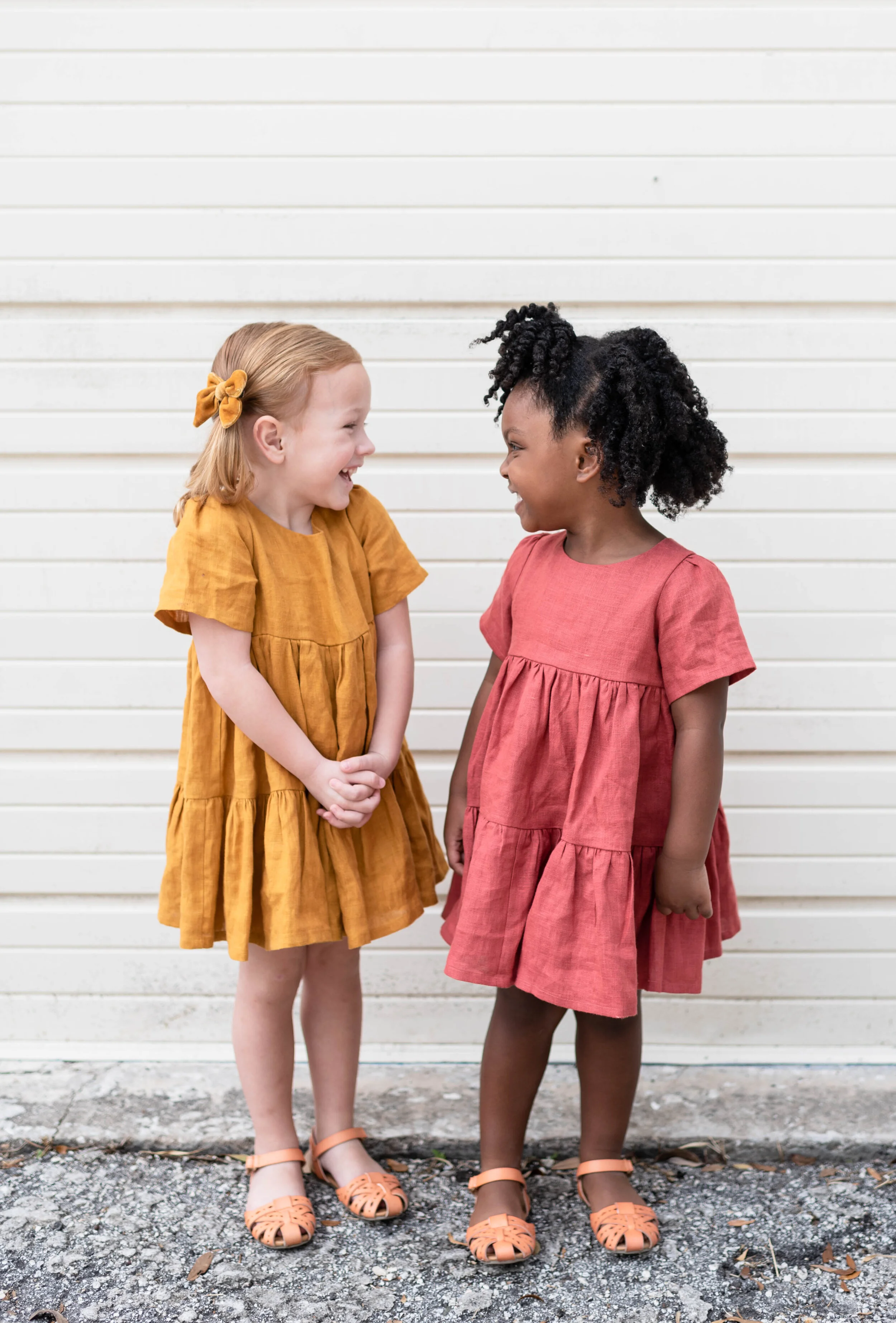 Two young girls in colorful dresses smiling and laughing at each other against a white wall.