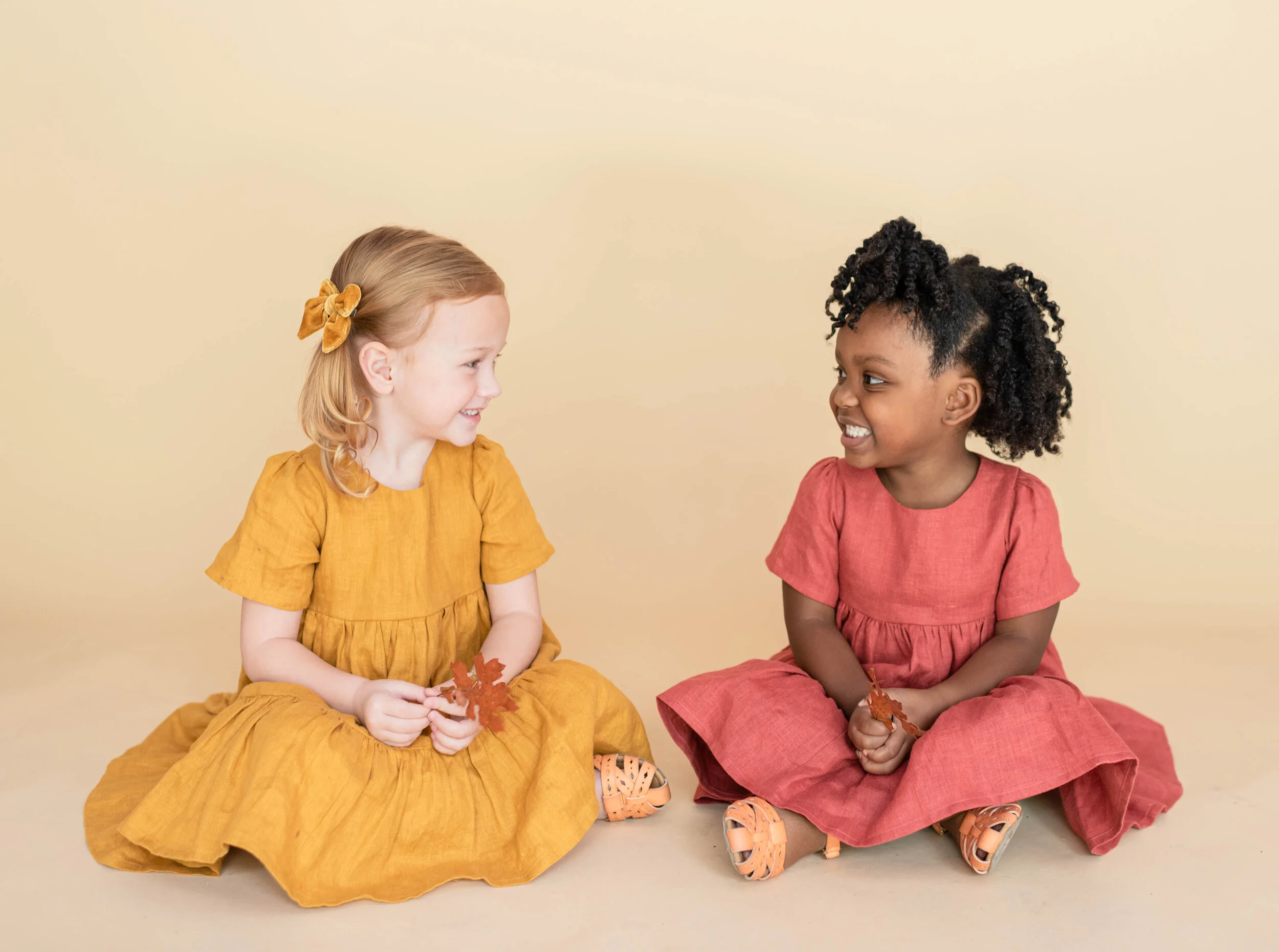 Two young girls sitting on the floor facing each other, smiling, holding autumn leaves. One girl is wearing a mustard yellow dress with a matching hair bow, and the other is dressed in a coral pink dress with orange sandals, against a plain beige bac